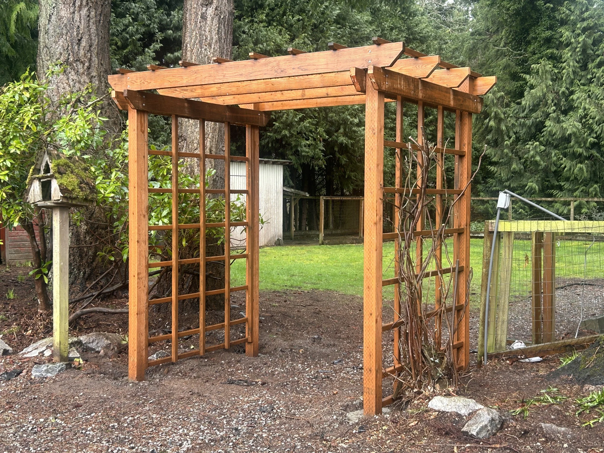 A wooden garden arbor structure with a pergola roof in a backyard surrounded by trees and greenery.