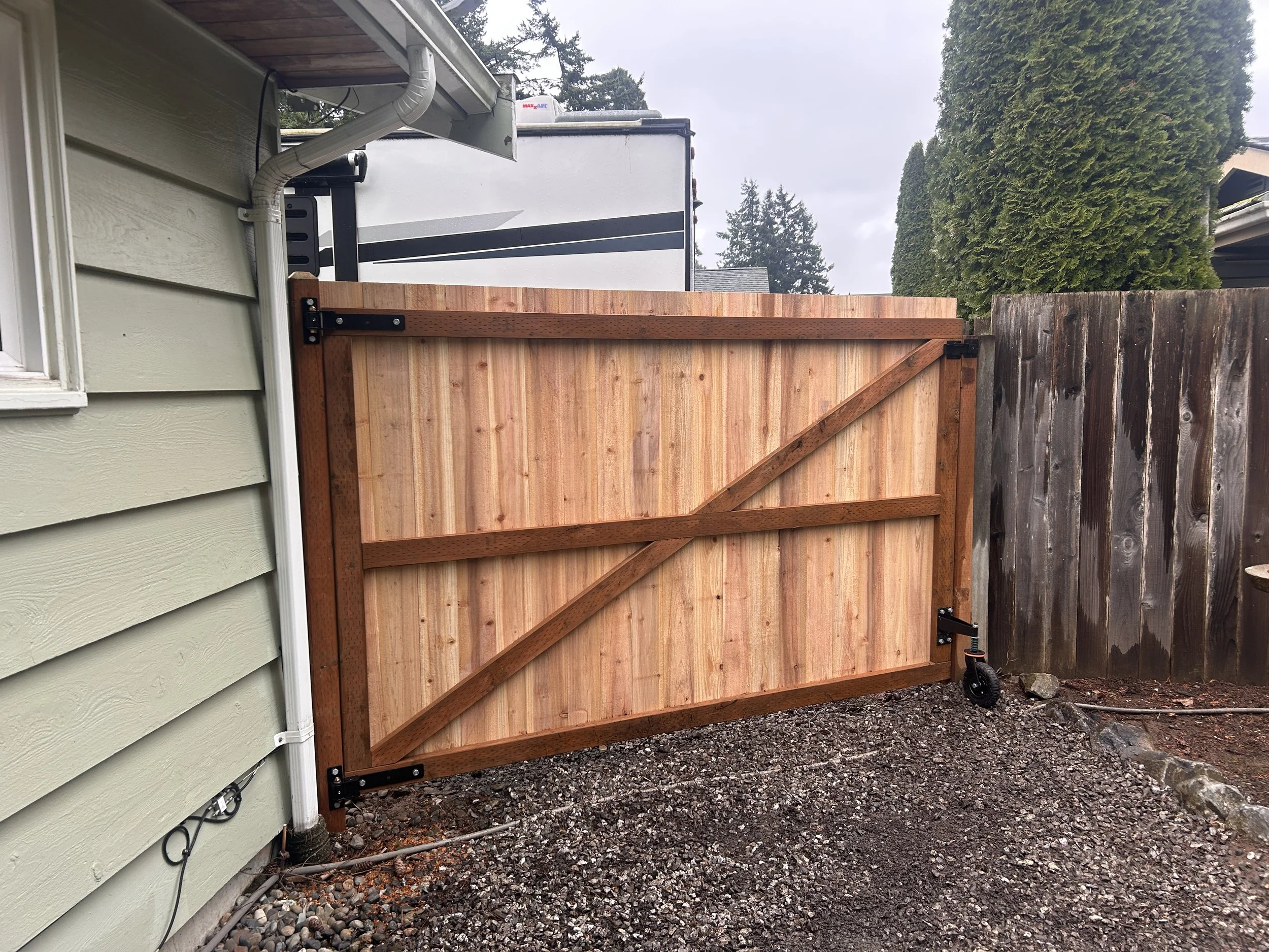 A newly installed wooden gate with metal brackets on a backyard fence, with a green house wall on the left and tall hedge on the right. The ground is covered with gravel and some rocks.