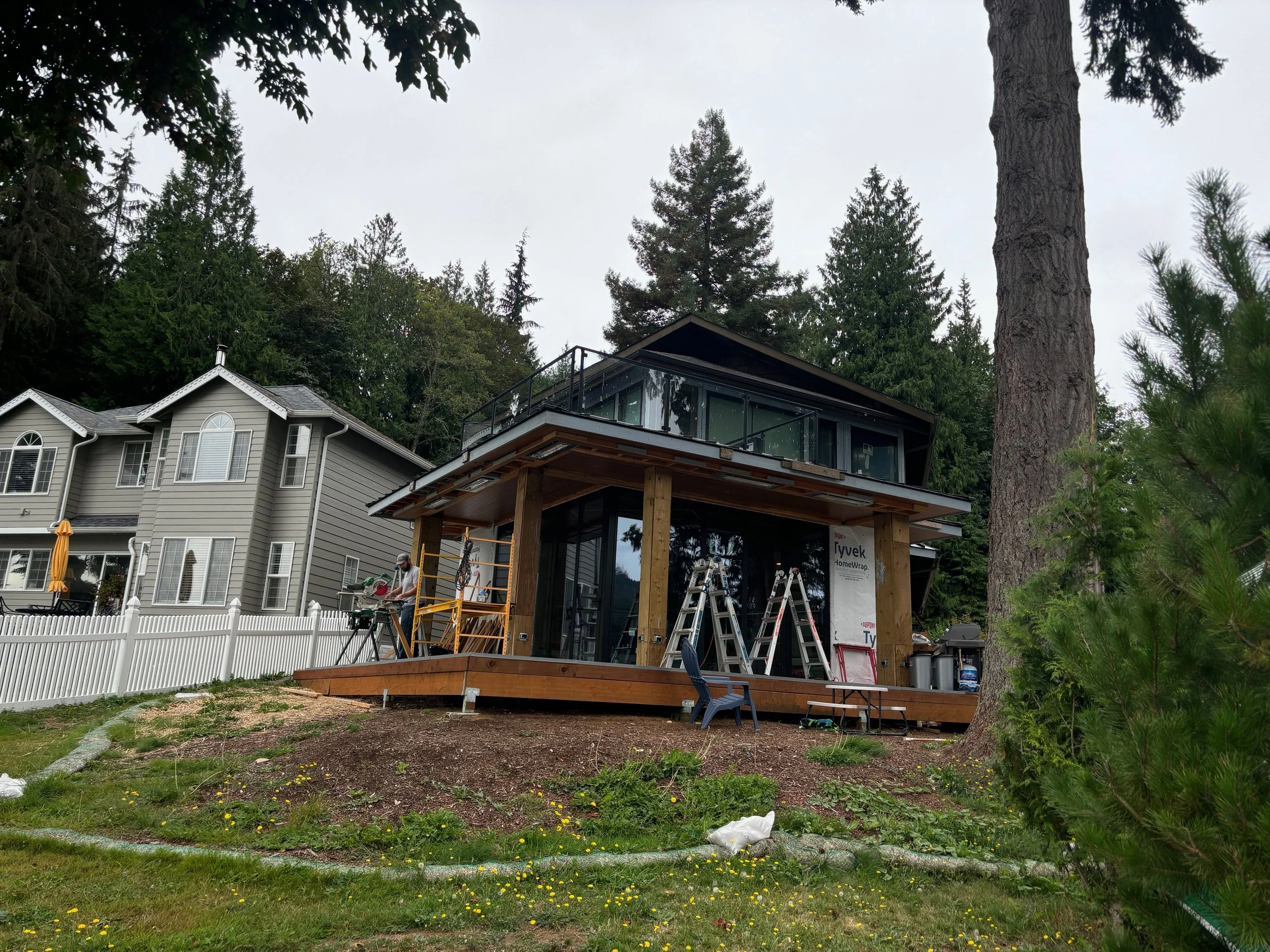 A house under construction with a modern glass addition, surrounded by tall trees and a white picket fence, with construction workers working and ladders nearby.