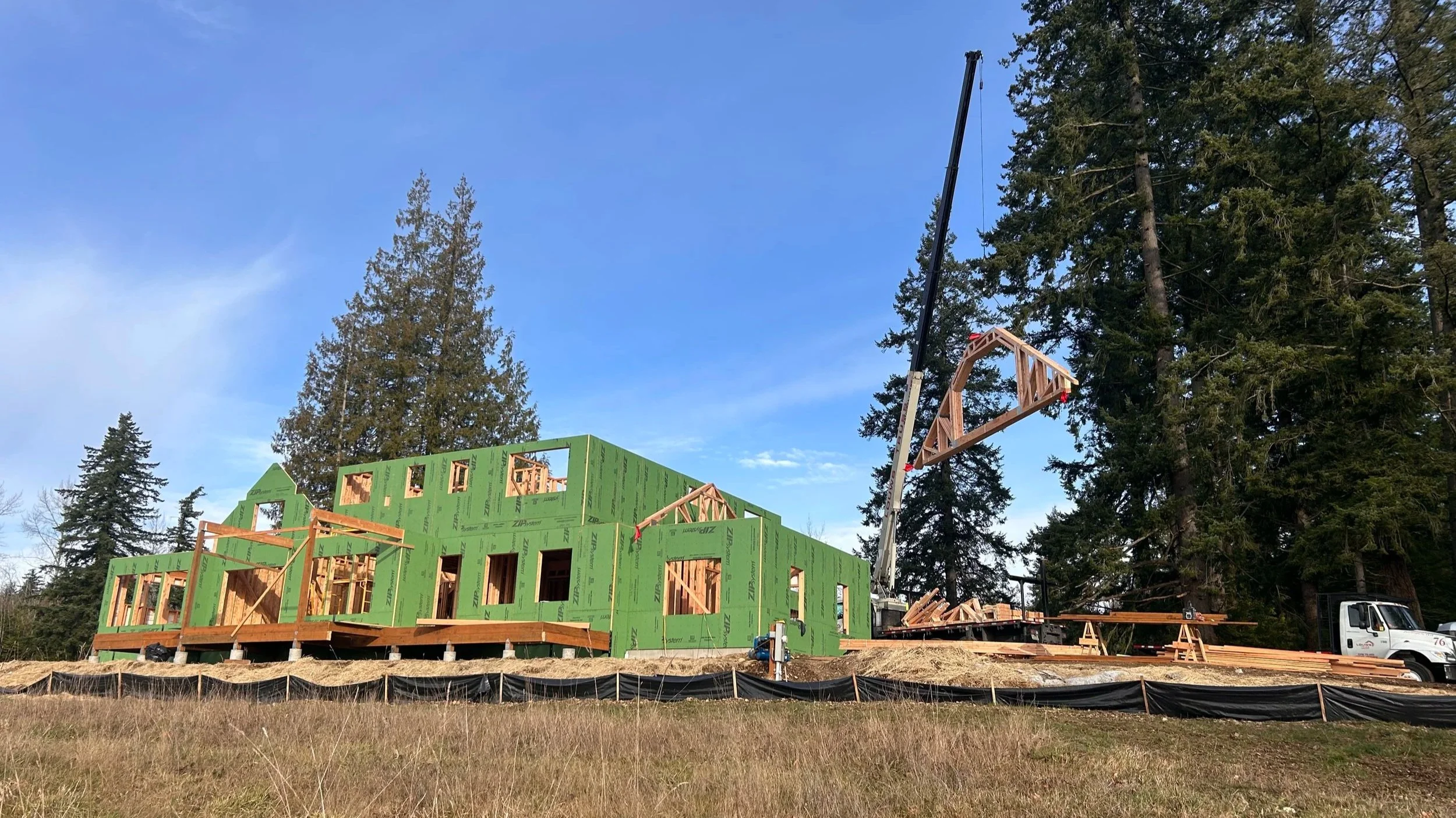 Construction site with a house in the framing stage, green insulation panels on the walls, wooden beams, a crane lifting a large wooden frame, and surrounded by tall trees under a blue sky.