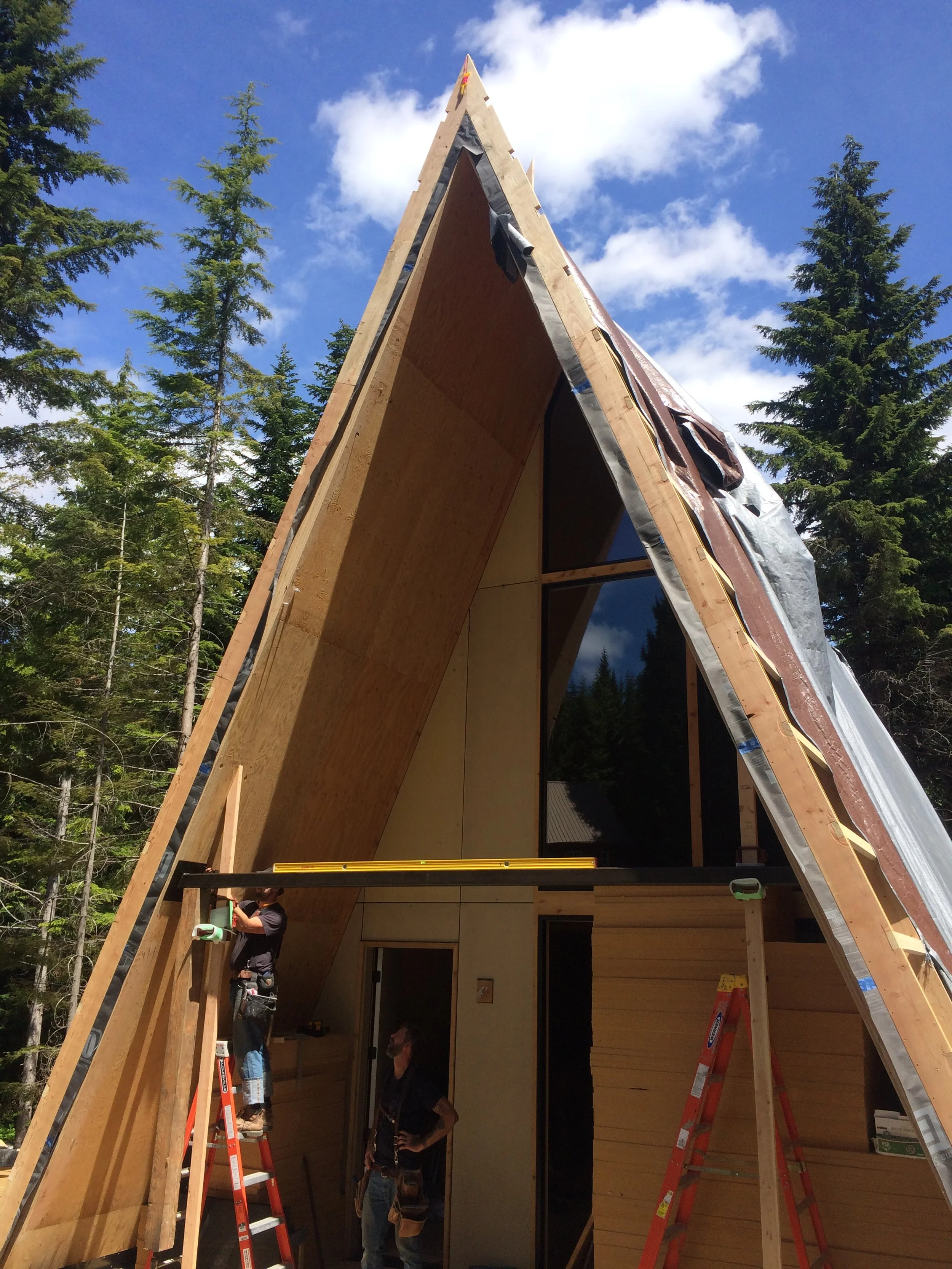 Construction workers building the A-frame structure of a house with wooden panels and insulation materials, surrounded by evergreen trees and a partly cloudy sky.