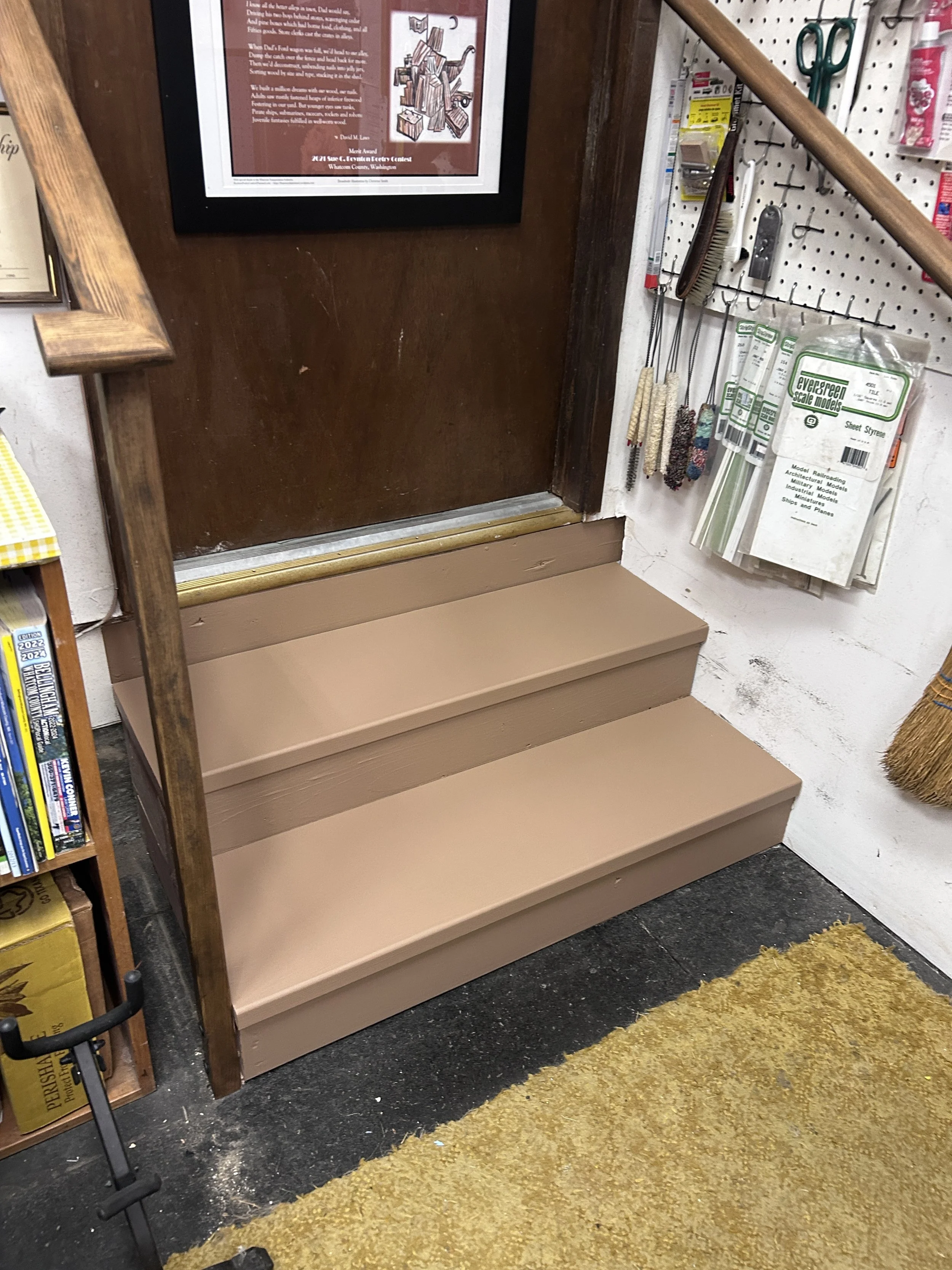 A set of three beige stairs leading up to a dark wooden door, with a gold-colored threshold. To the right of the stairs, there is a white wall with various tools and items hanging on a pegboard, including brushes, scissors, and packaged model train s
