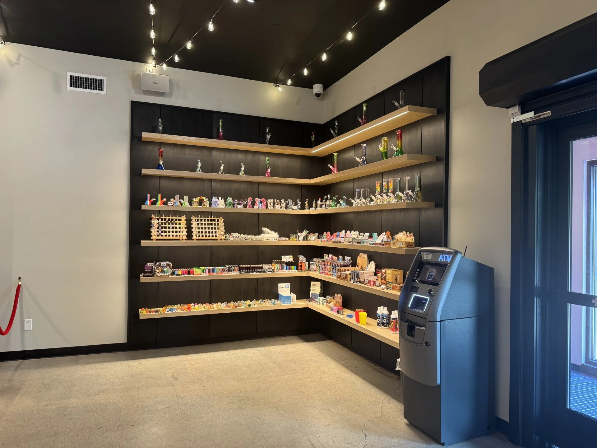 Store wall with shelves displaying glass bongs and smoking accessories, next to an ATM machine, with a security camera and ceiling lights.