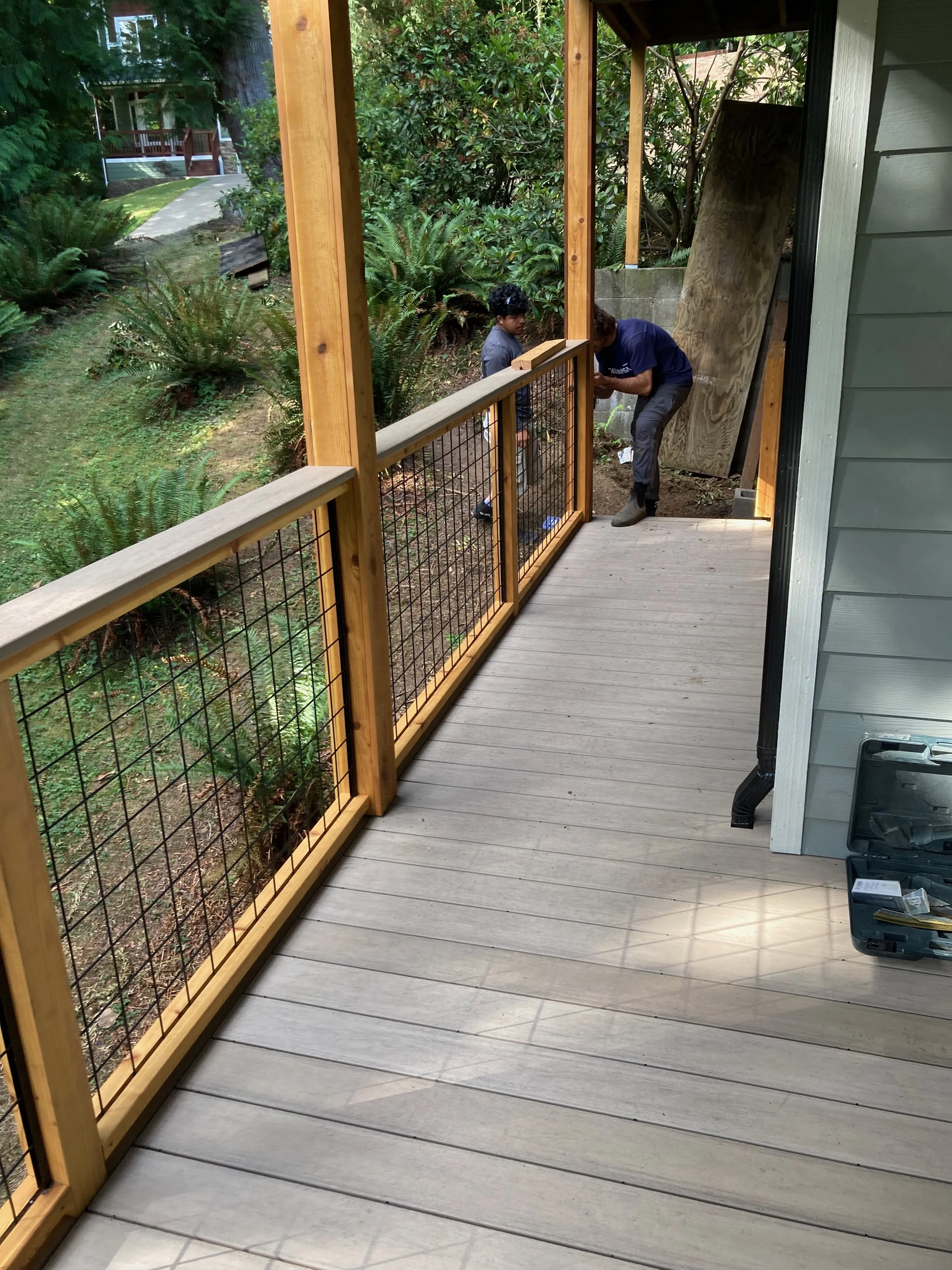 People installing a wooden and wire fence on a balcony next to a green yard with plants and trees.