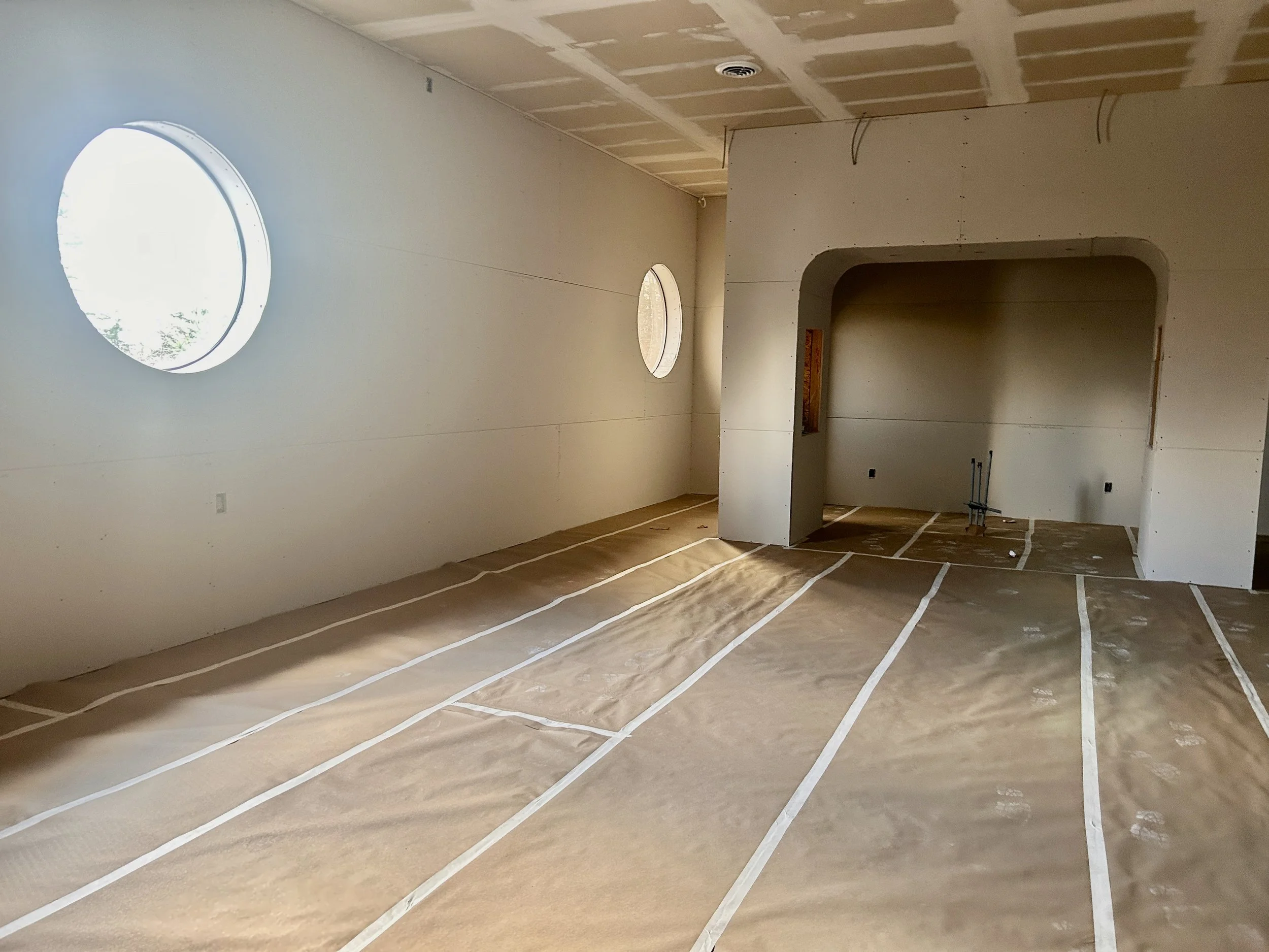 Interior view of a house under construction with drywall installed. The ceiling has sections and circular openings for lights or vents. The floor is covered with paper or protective material, and some electrical wiring is visible.
