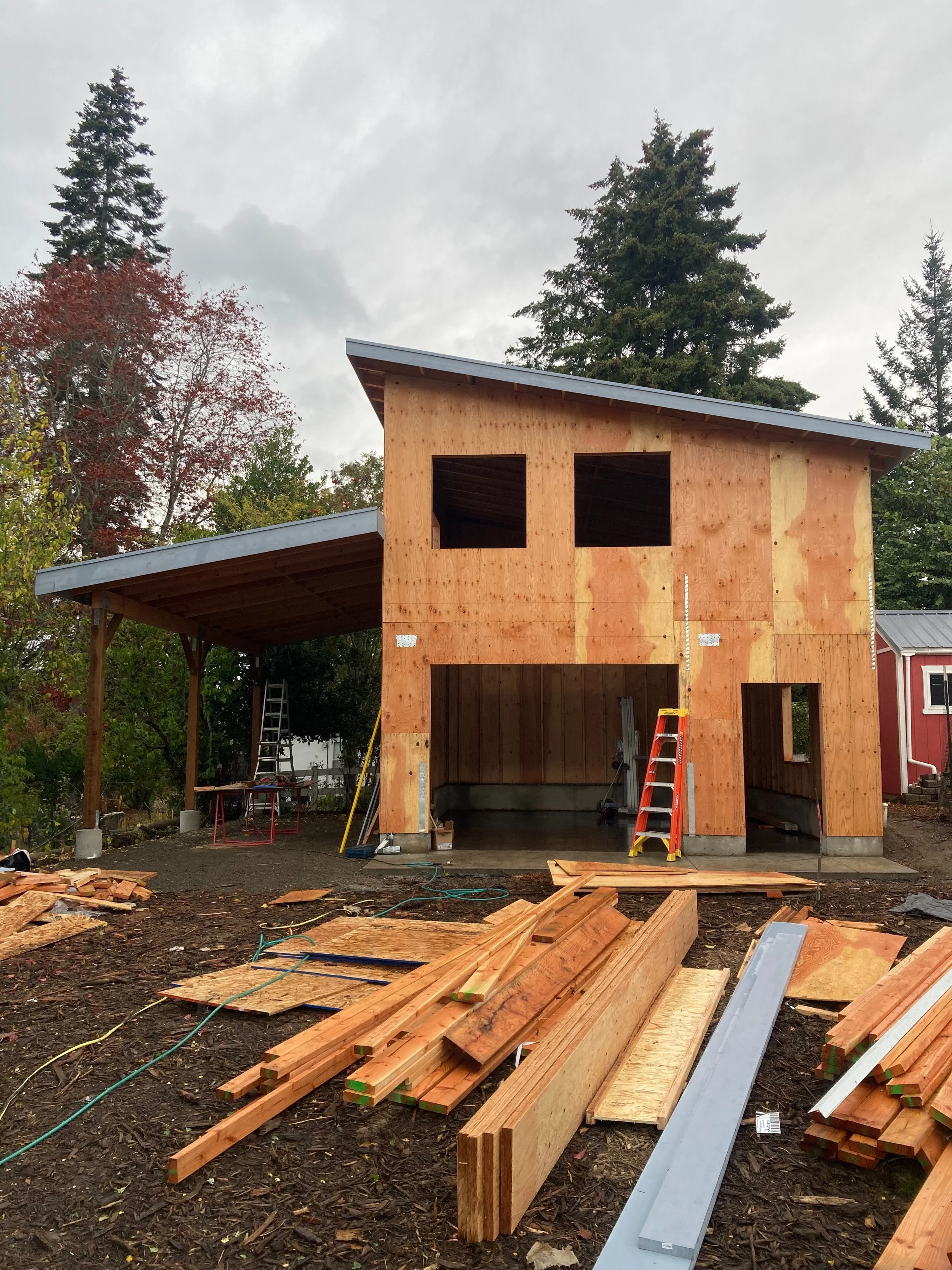 Construction site with a partially built wooden house, construction tools, and building materials scattered on the ground, surrounded by trees and cloudy sky.