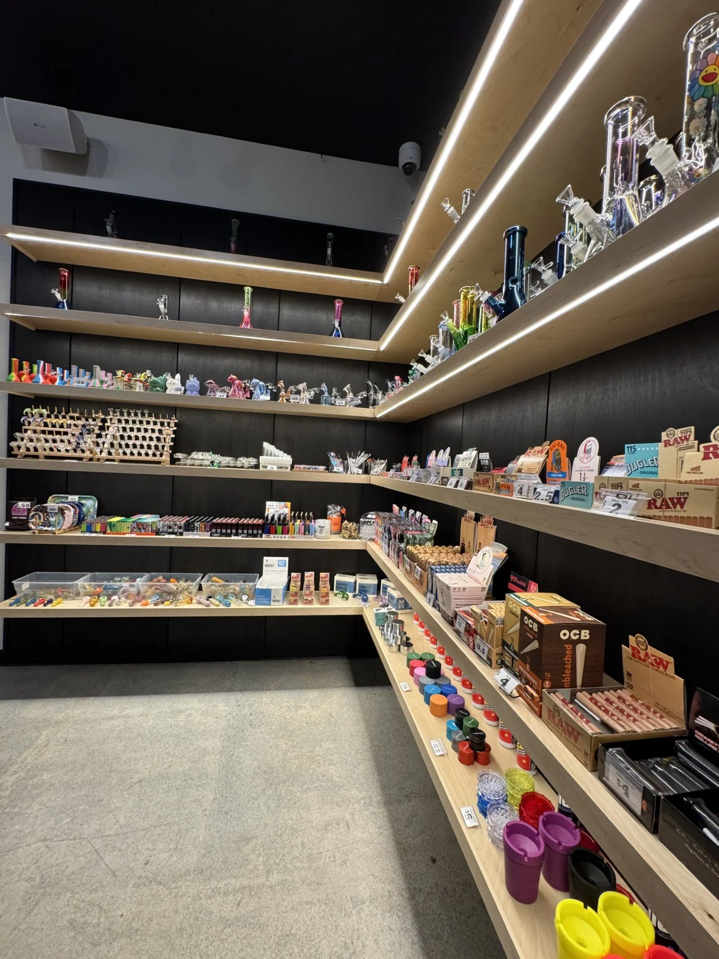 A retail store display with shelves stocked with smoking accessories and smoking-related products such as glass pipes, vaporizers, grinders, rolling papers, lighters, and storage containers.