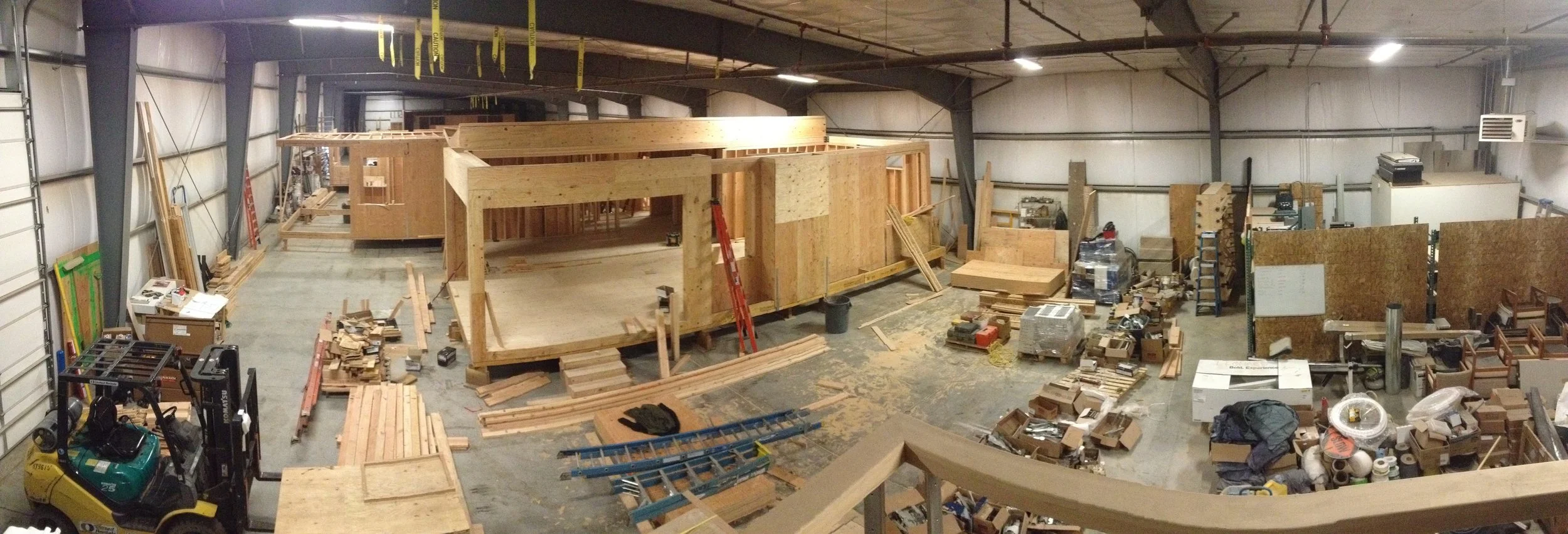 Construction warehouse with wooden framing in progress, scattered tools, lumber, and building supplies, and a forklift in the foreground.