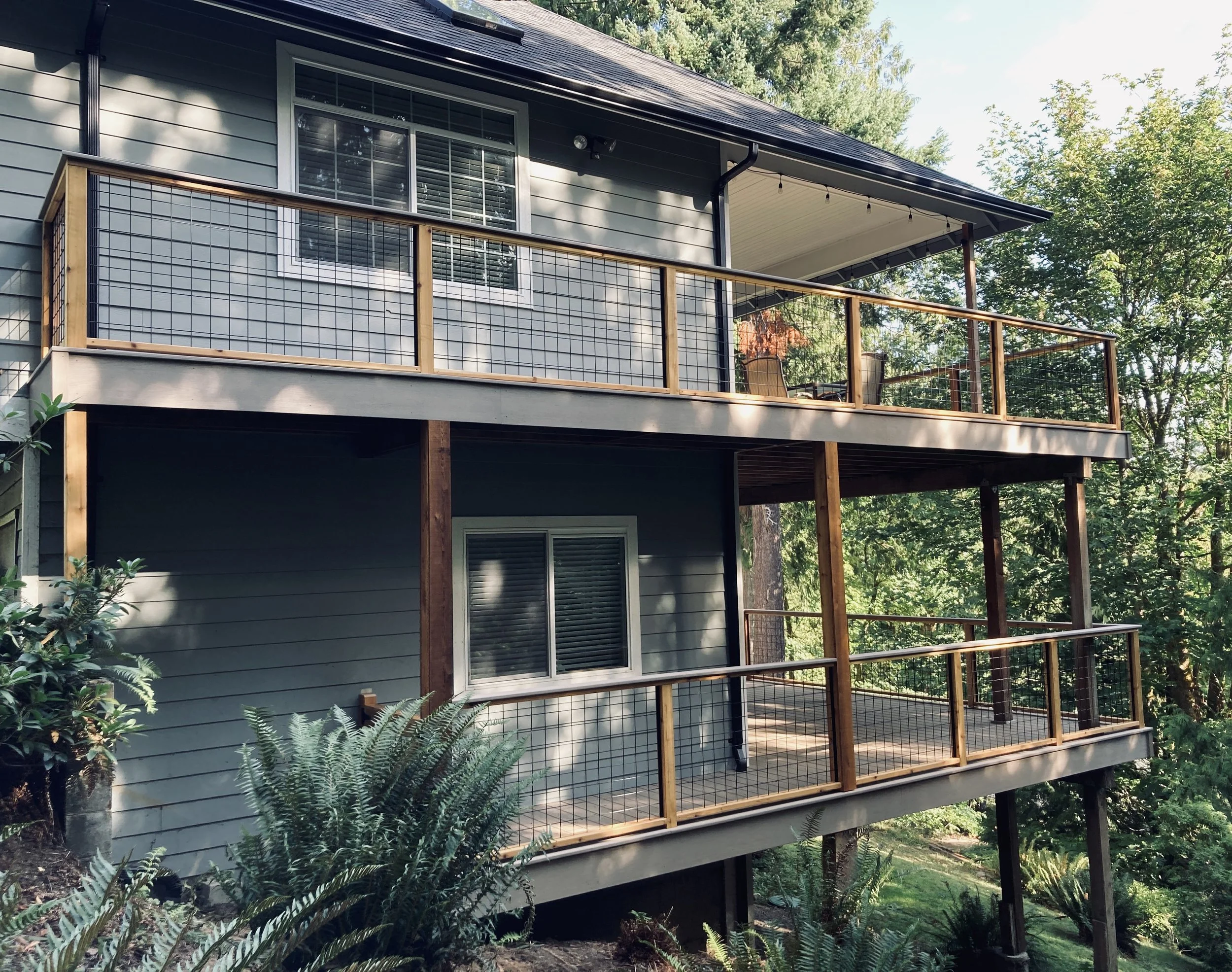 Two-story house with light gray siding, large windows with white frames, a balcony with wire fencing and wooden posts, and surrounded by trees and greenery.