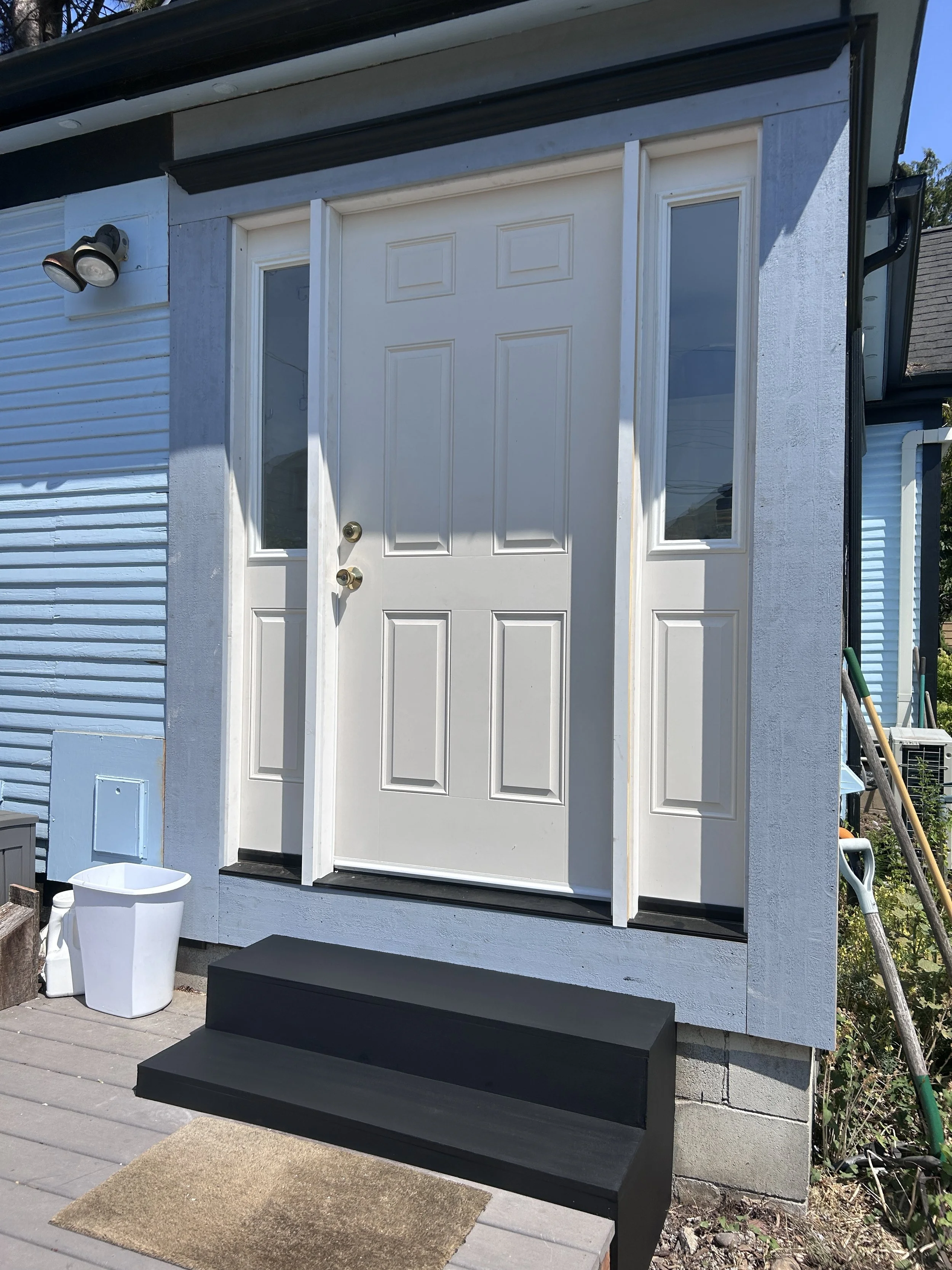 Front entrance of a house with white door, two narrow windows on each side, black steps, and a tan doormat in front. There is a white trash bin to the left of the door and a light fixture with two bulbs mounted on the siding. Gardening tools and an a