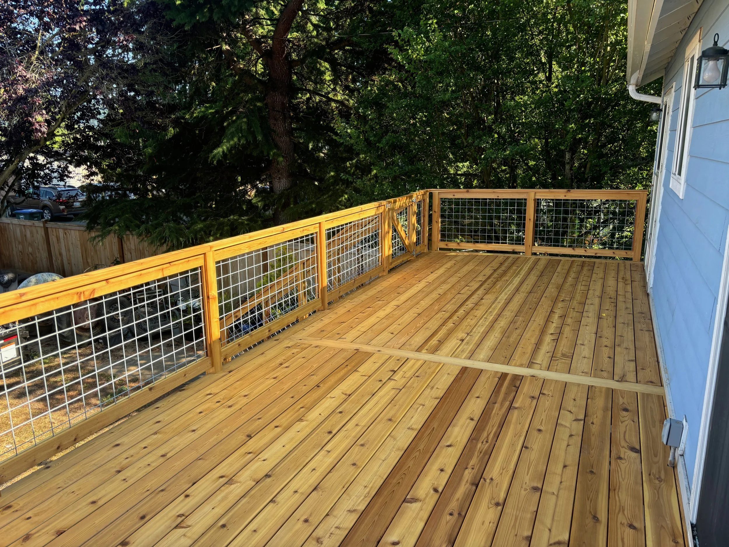 A wooden deck with a railing attached to a house, overlooking a backyard dense with trees, and a row of parked cars visible behind the fence.