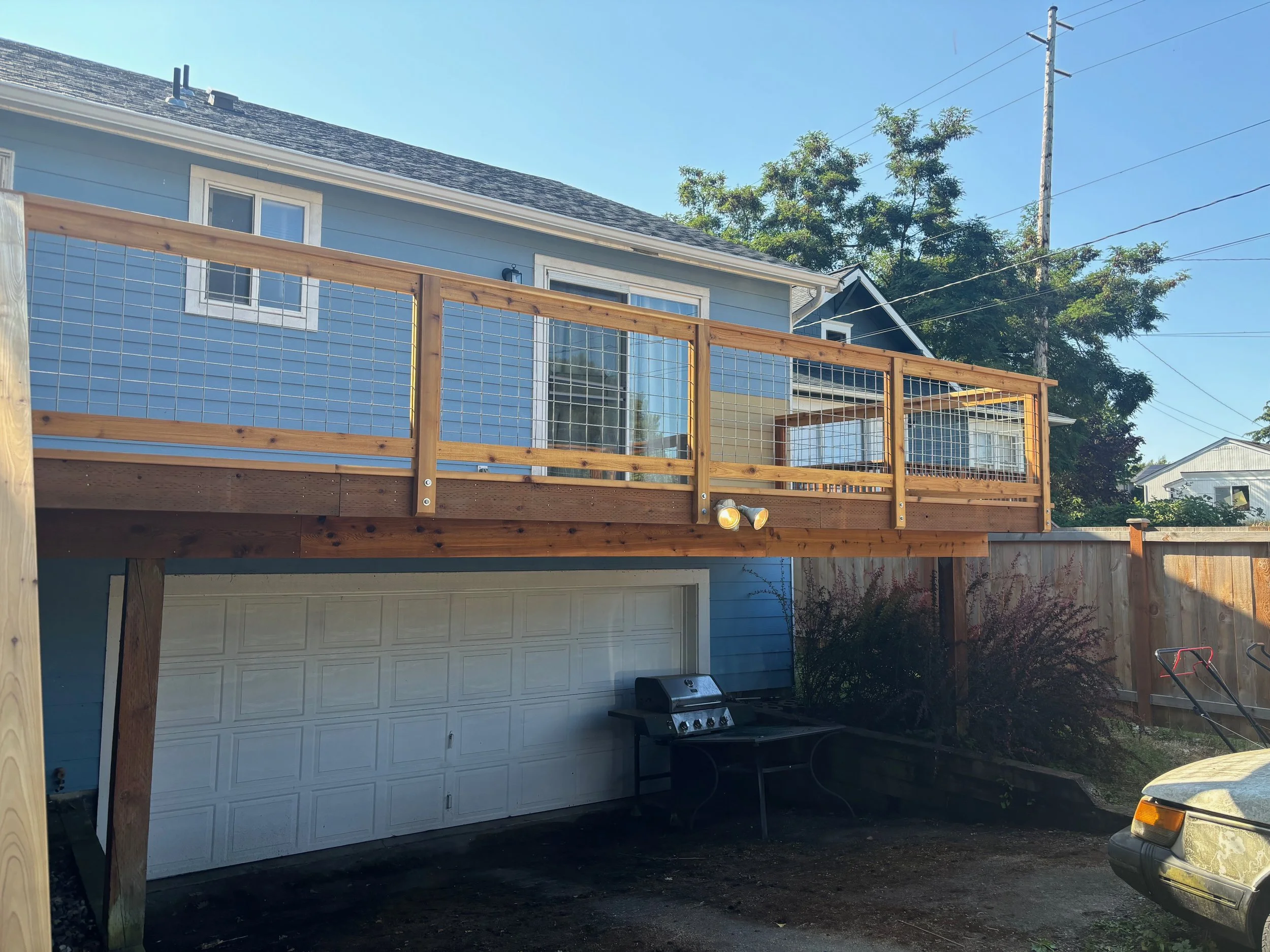 The image shows an exterior view of a light blue house with a large backyard deck made of wood with metal wire railing. There is a white garage door underneath the deck, and a barbecue grill on the ground in front of the garage. A garden hose is visi