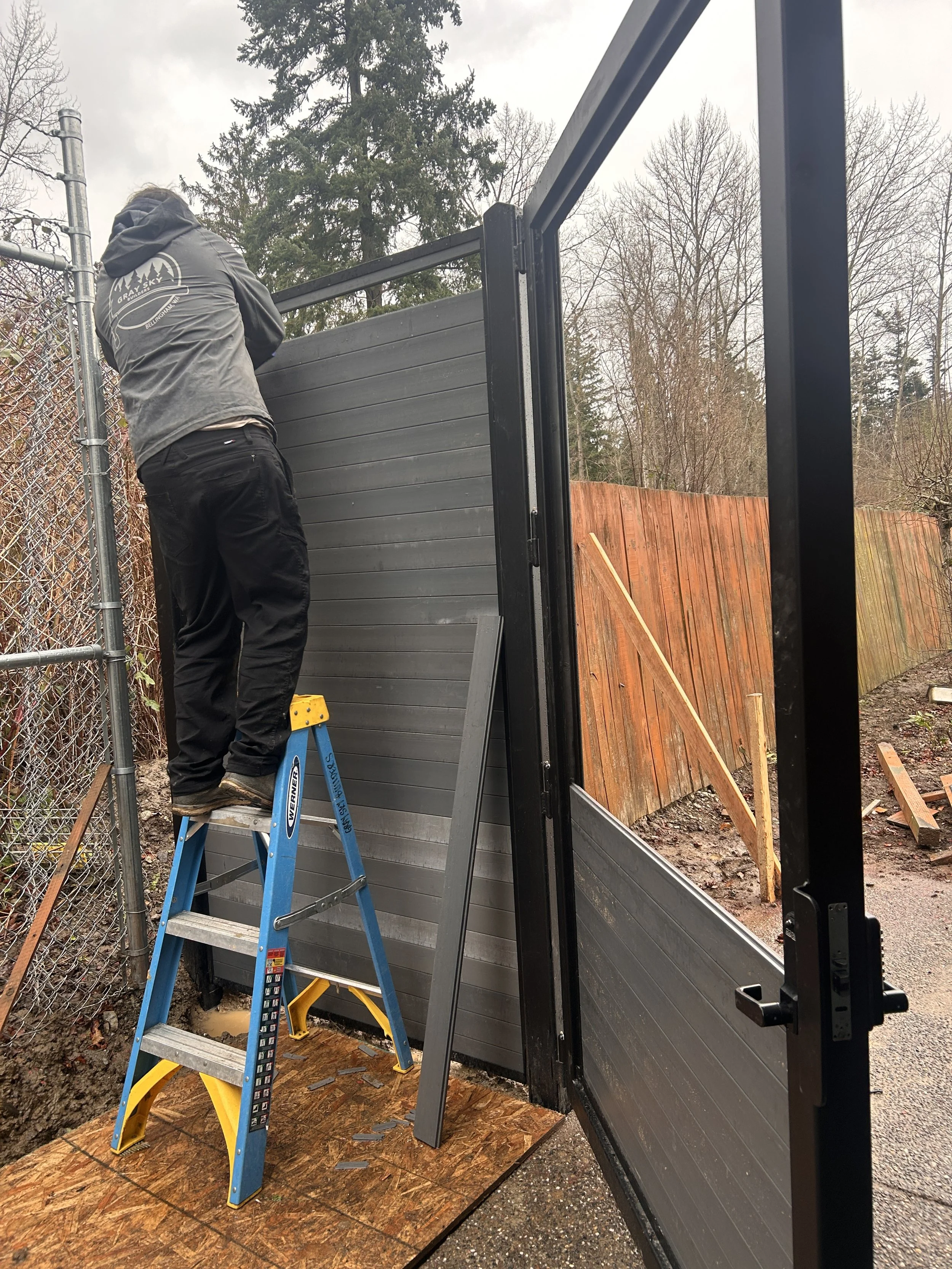 A person working on a black fence panel outdoors during daytime, standing on a blue and yellow step ladder, with trees and a wooden fence in the background.