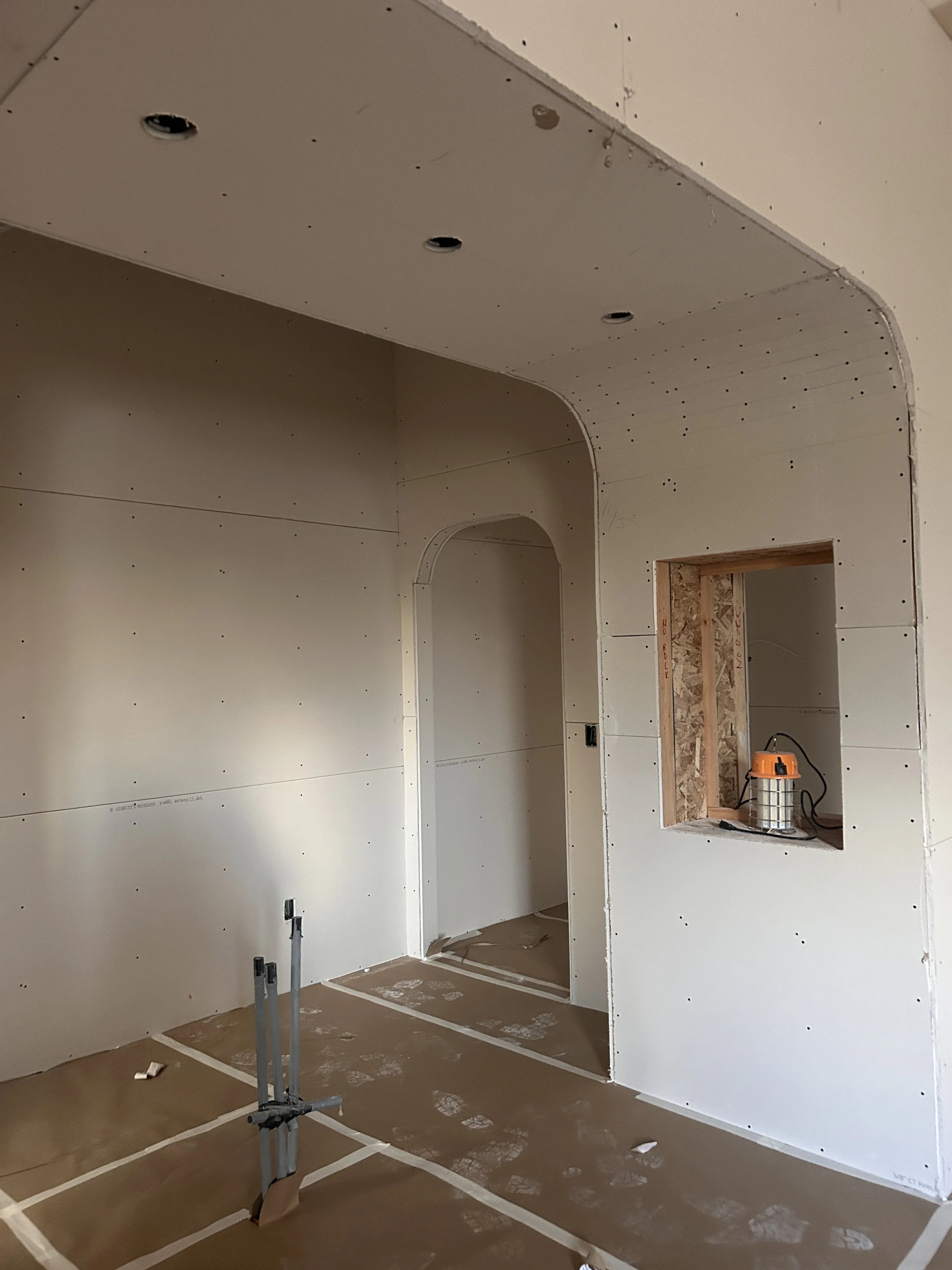 Interior of a room under construction with drywall walls and ceiling, electrical outlets and a small built-in shelf. Construction tools are present on the floor.