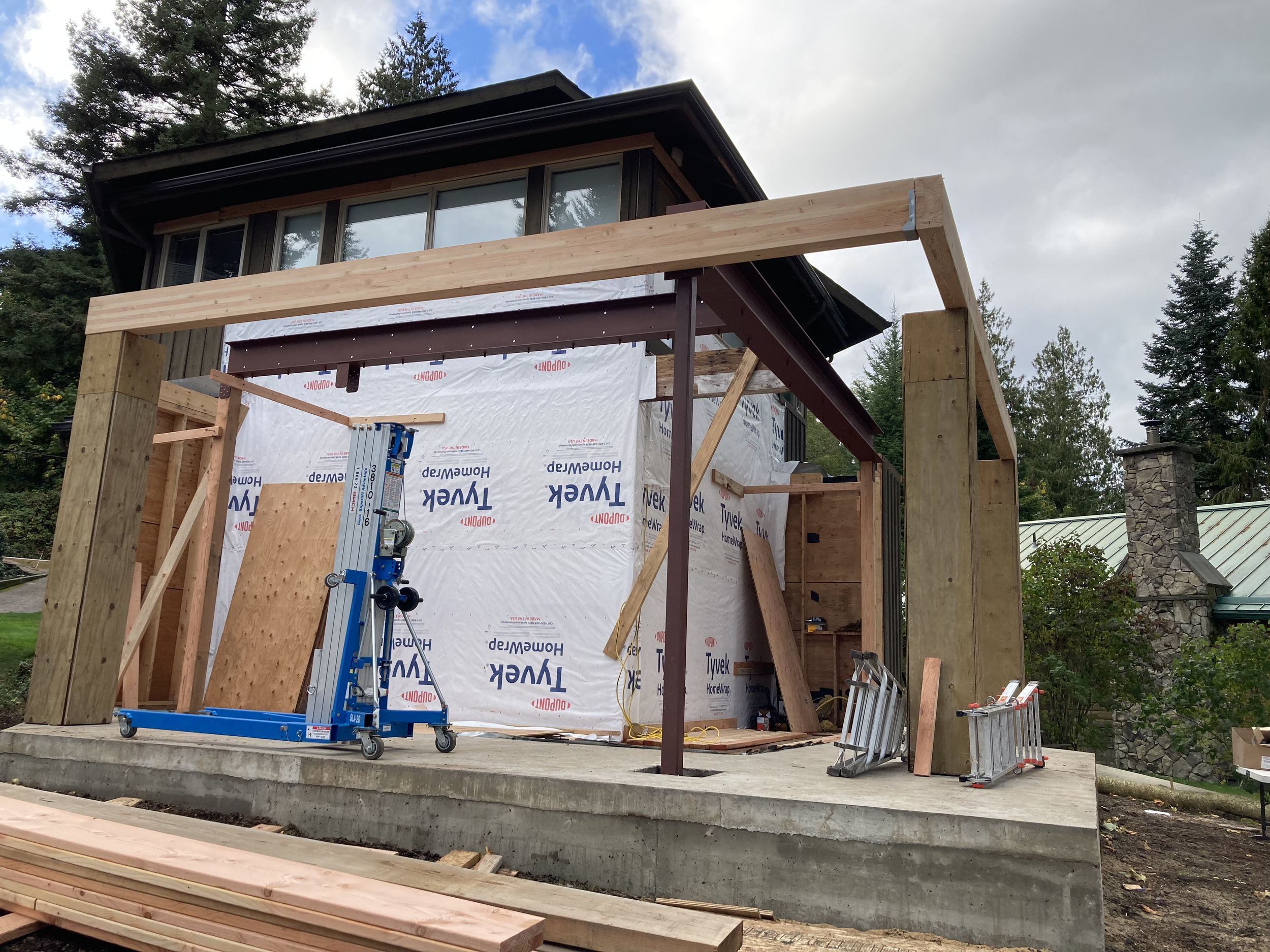 Construction site of a house with wooden framing, scaffolding, construction tools, and a partially built elevated porch or deck. Trees and a cloudy sky are visible in the background.
