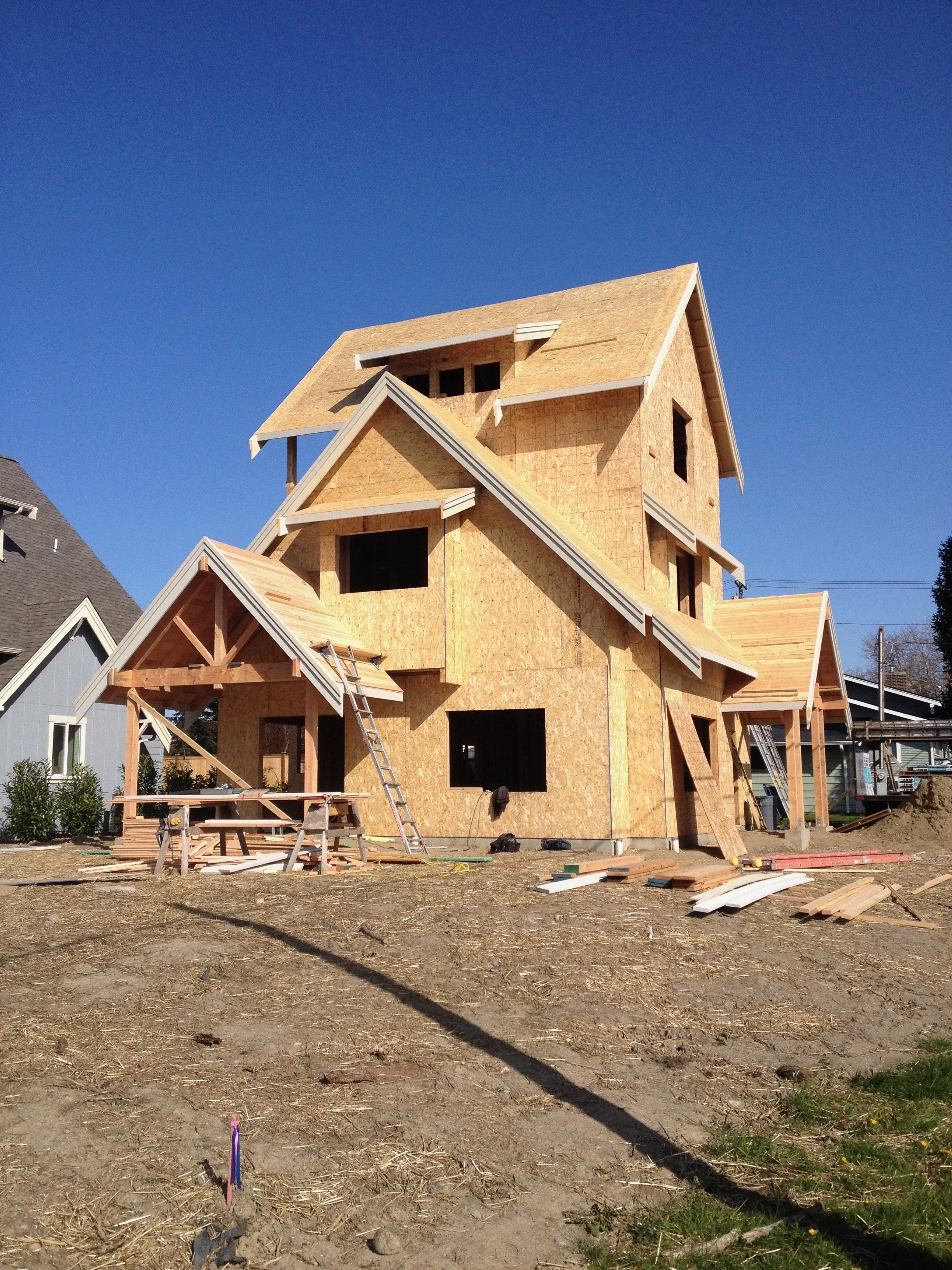 A house under construction with plywood walls and multiple gabled roofs, set against a clear blue sky, with construction tools and materials scattered on the ground in front.