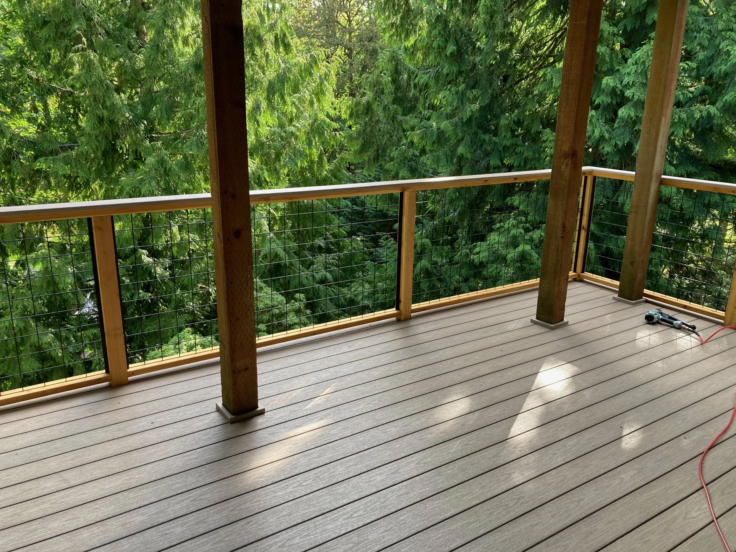 A wooden balcony with safety wire railing overlooking lush green trees.