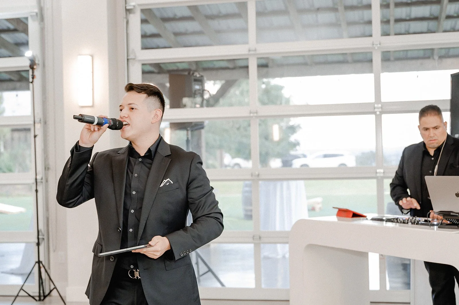 A man in a black suit singing into a microphone at an indoor event, with a DJ or sound technician working at a nearby table with a laptop and DJ equipment in the background.