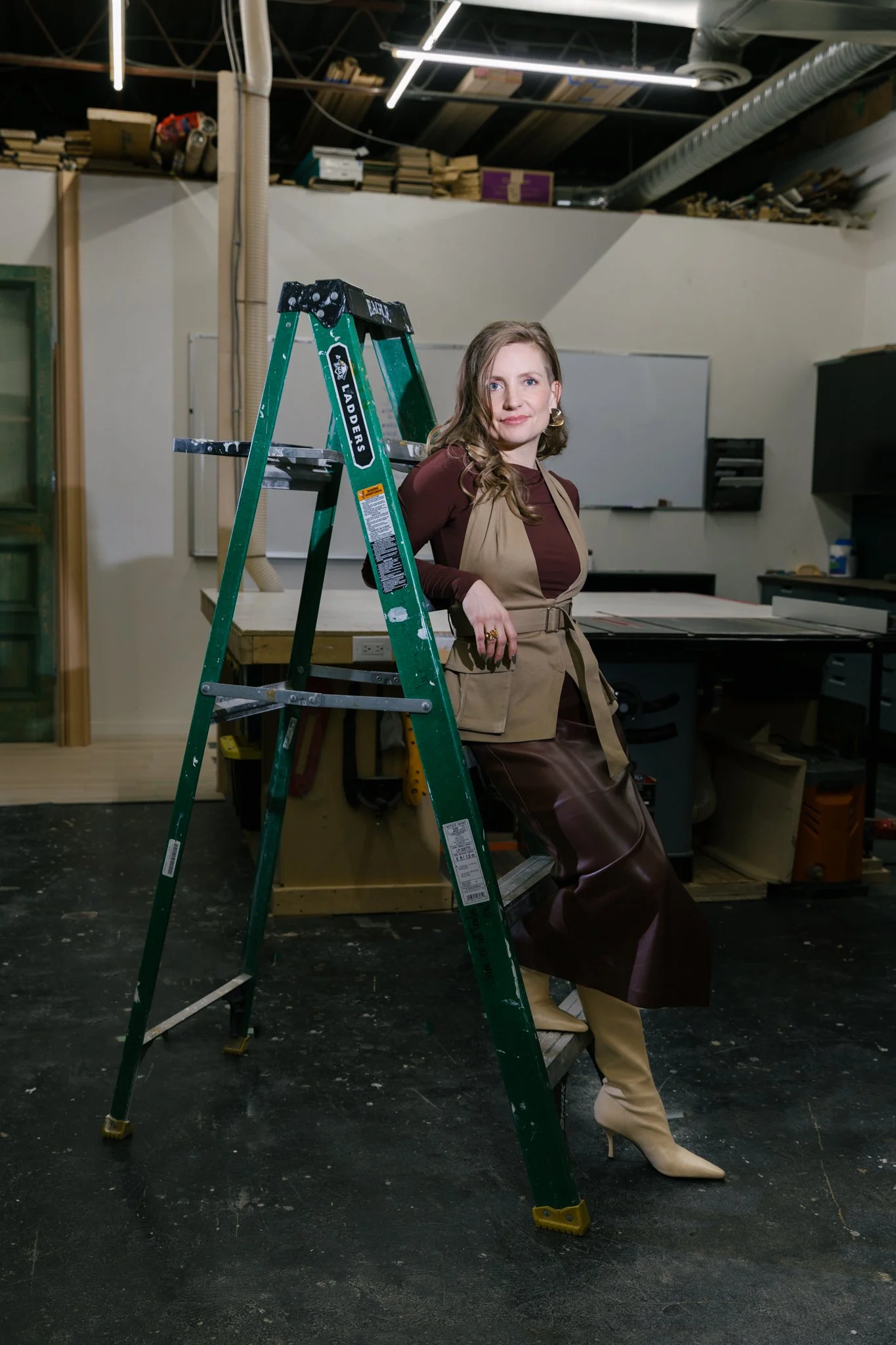 A white woman leaning on a green ladder in a workshop dressed fashionably in a brown leather skirt, white leather boots, and brown shirt with a beige vest looking confidently at the camera
