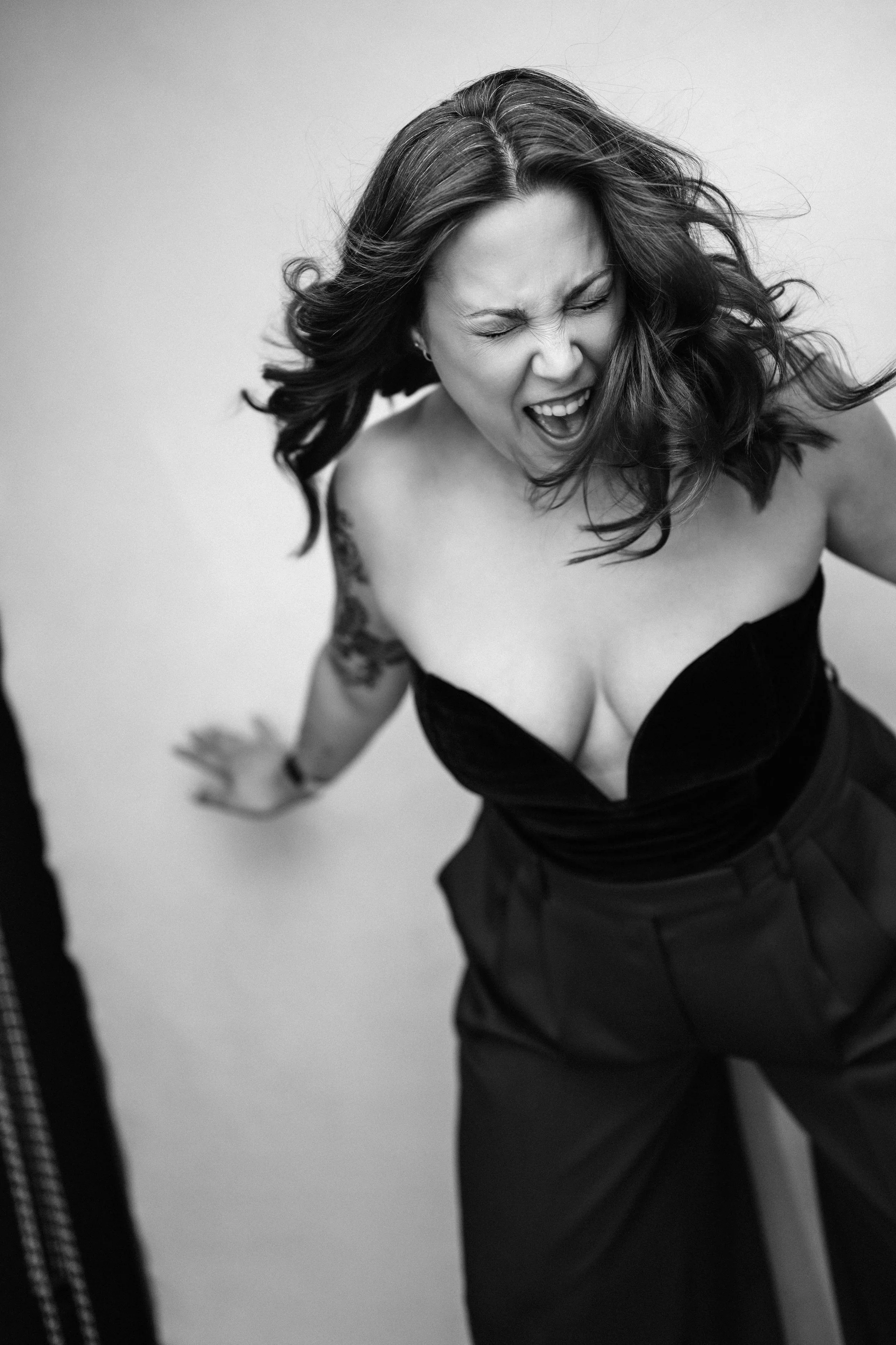 A black and white image of a woman screaming while a fan blows her hair around, her arms flung behind her.