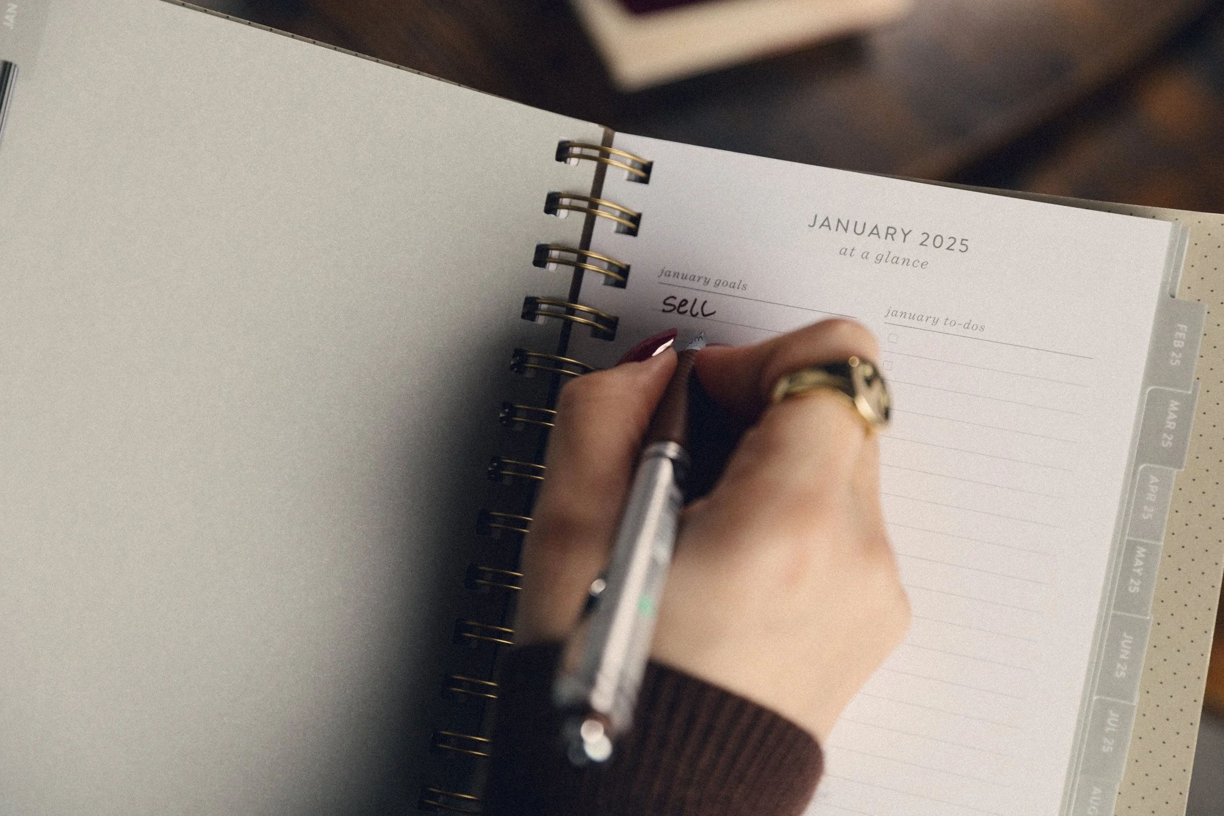 A woman's hand with burgundy nails and a gold ring holding a pen to a notebook calendar writing "sell" on the page
