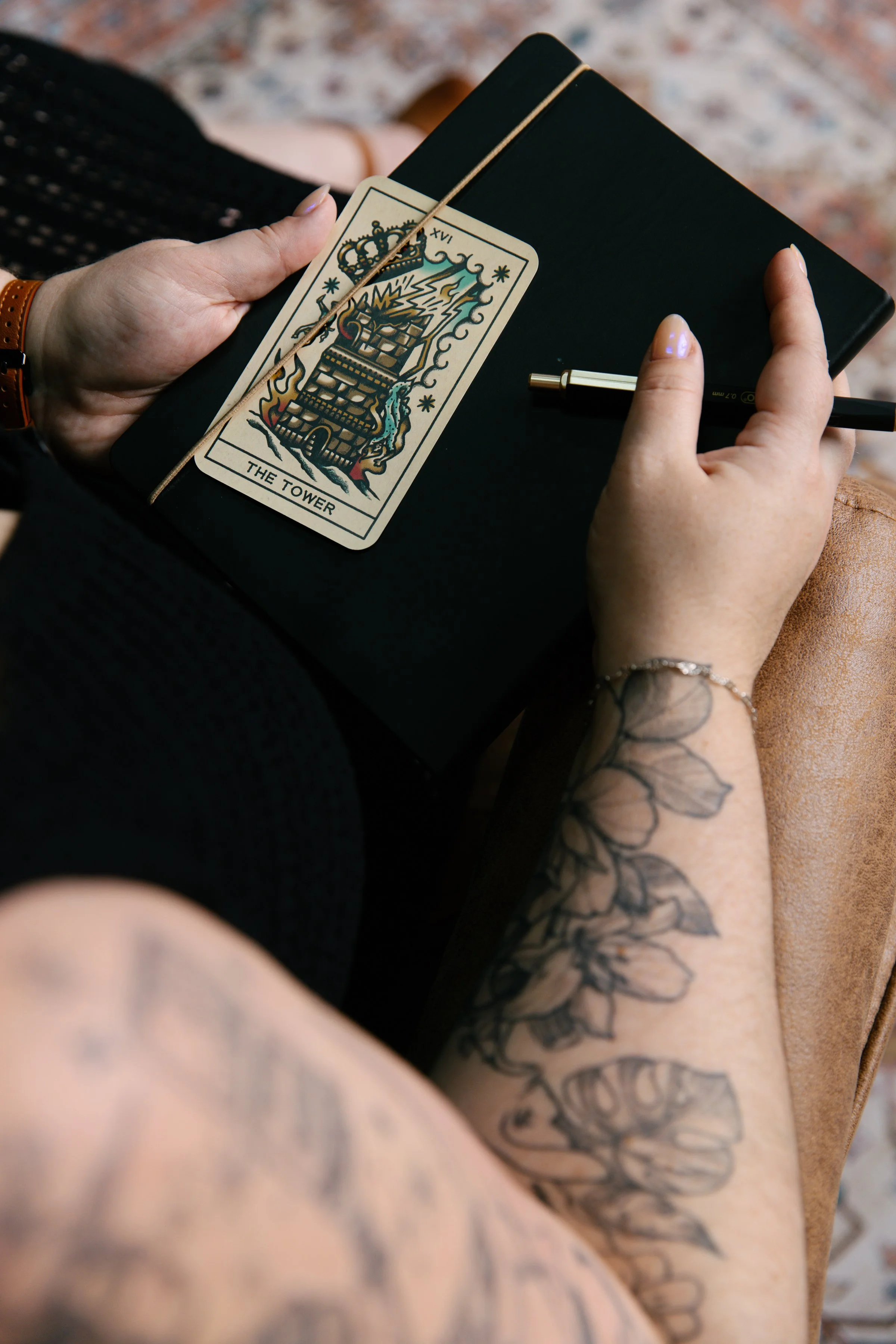 A women's floral tattooed arm holding a black notebook with The Tower tarot card on top