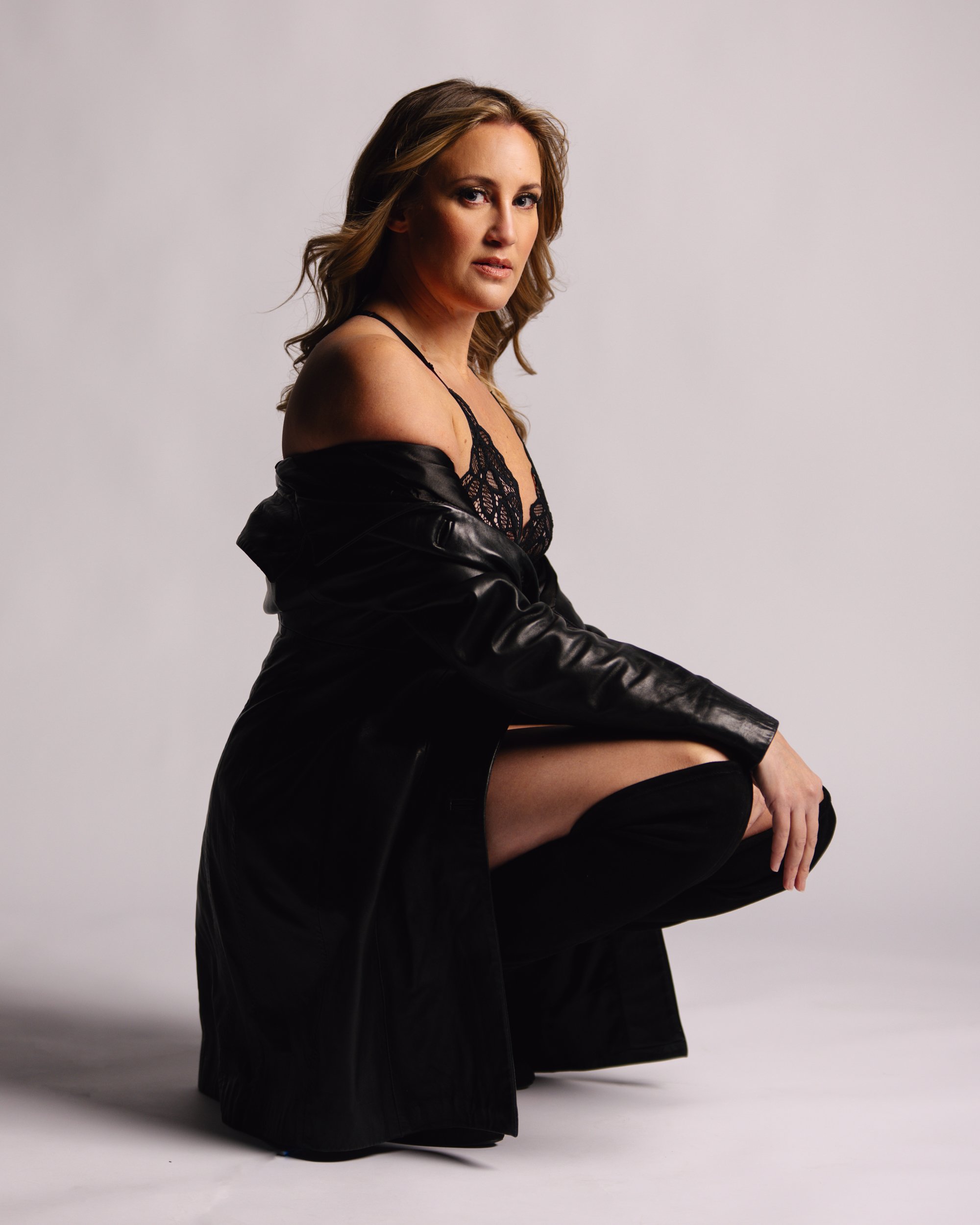 A woman with wavy blonde hair wearing a black lace lingerie, a partially off black leather jacket, and black thigh-high boots, posing against a plain grey background.