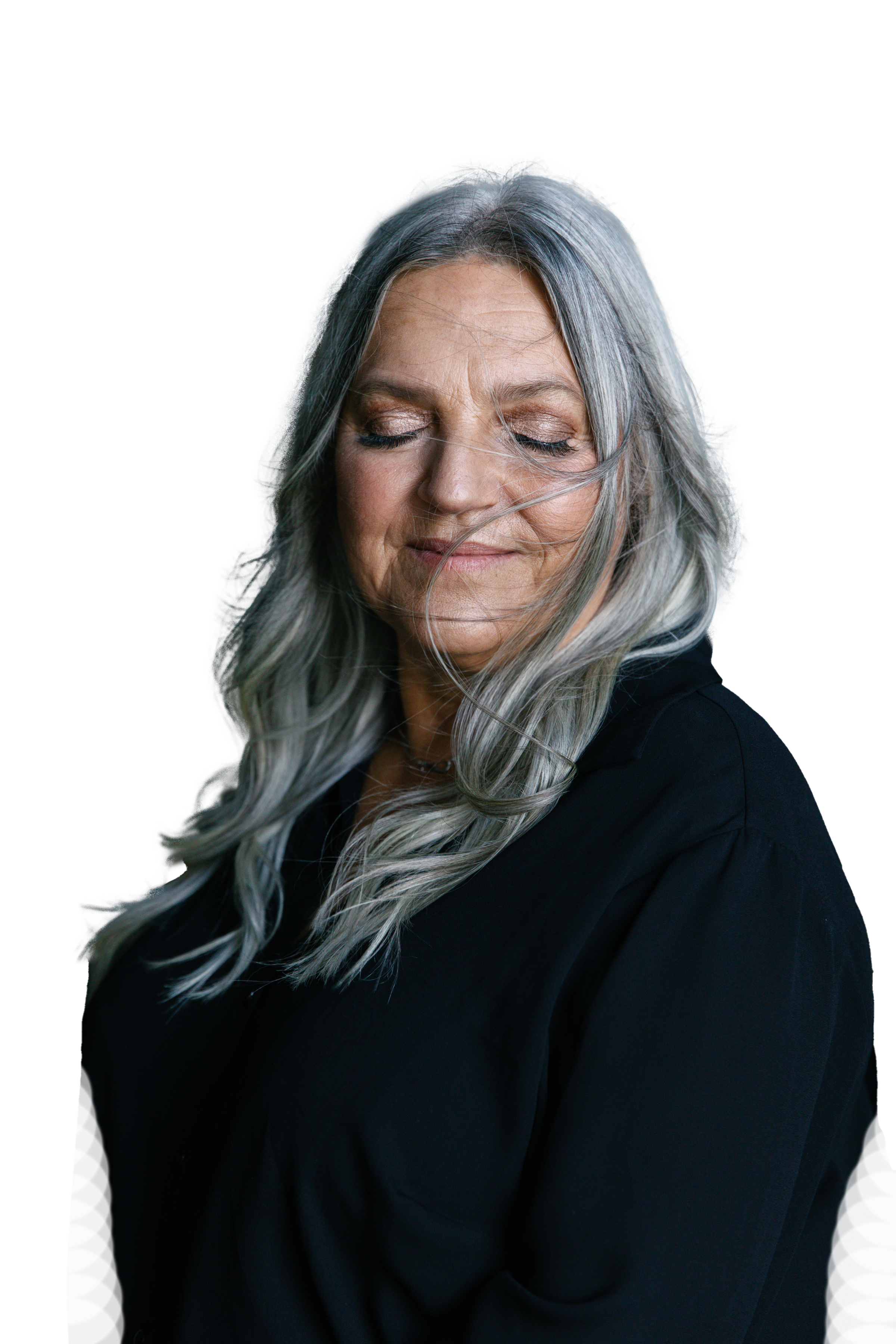 A woman with long gray hair, wearing a black top, smiling with her eyes closed against a dark background.