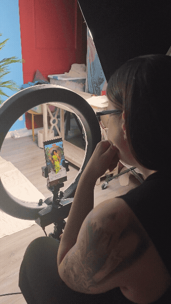 Person recording a video of a colorful portrait on a phone with a ring light setup in a room with red walls and a blue door.