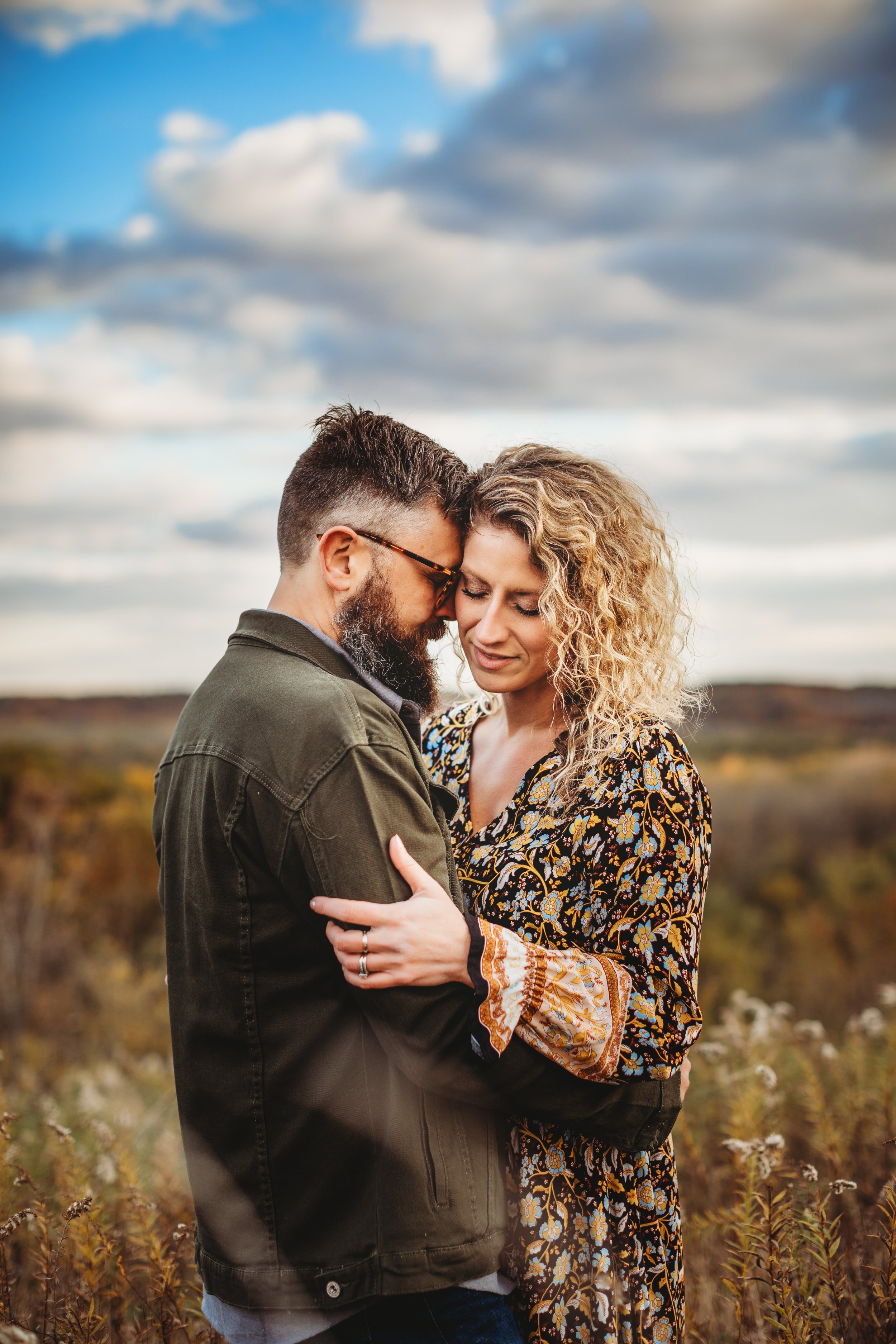  Under a blue sky, a husband and wife hug one another during an unposed family portrait session with Teala Ward Photography. parents #tealawardphotography #tealawardfamilies #springvalleyillinois #springvalleyphotographers #Illinoisfamilyphotographer