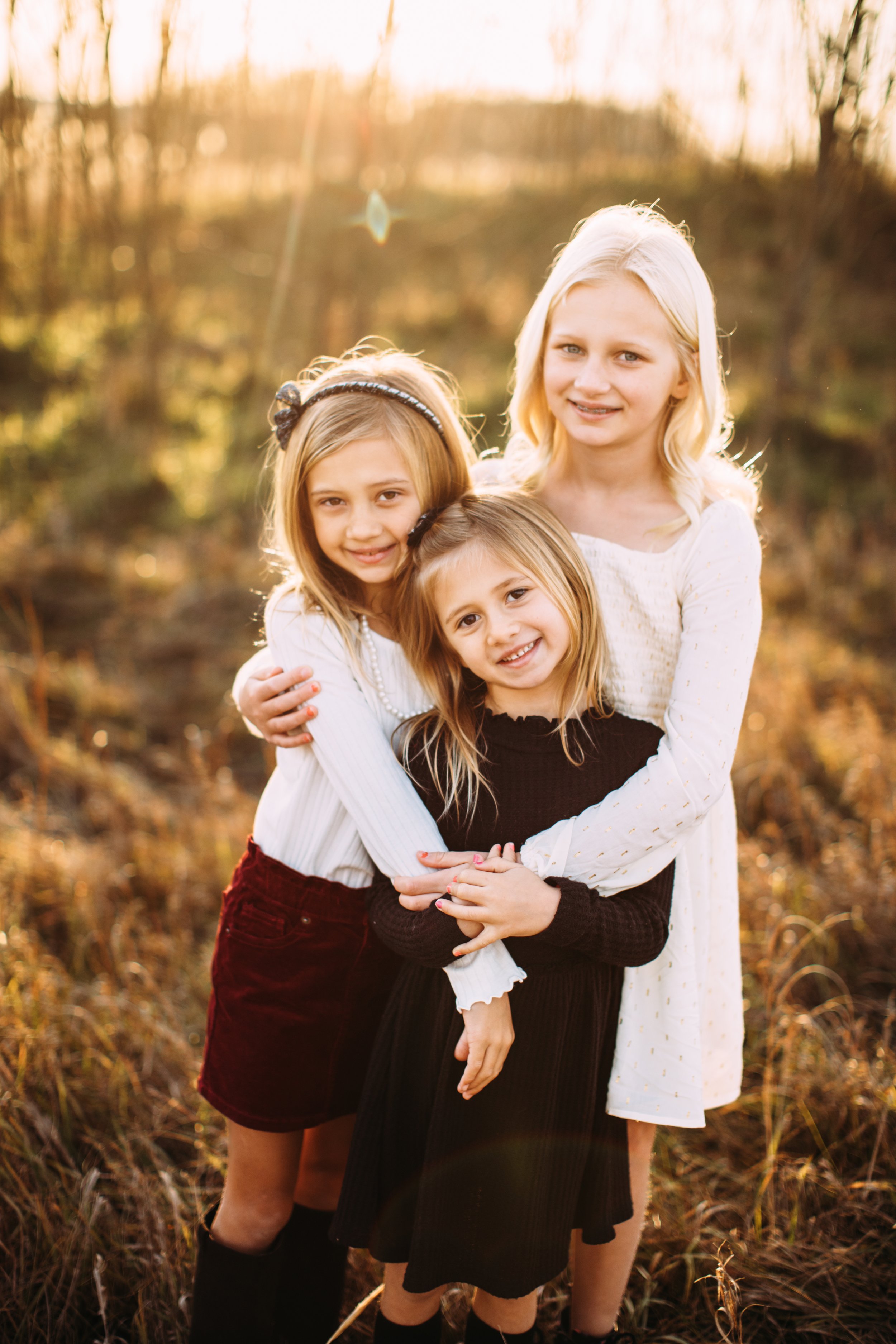  With the sun setting behind them, three young sisters hug in a yellow grass field by Teala Ward Photography. sisters sunset grass field #TealaWardPhotography #TealaWardFamilies #StarvedRockStatePark #BuffaloRockPhotographers #UticaIllinoisphotograph