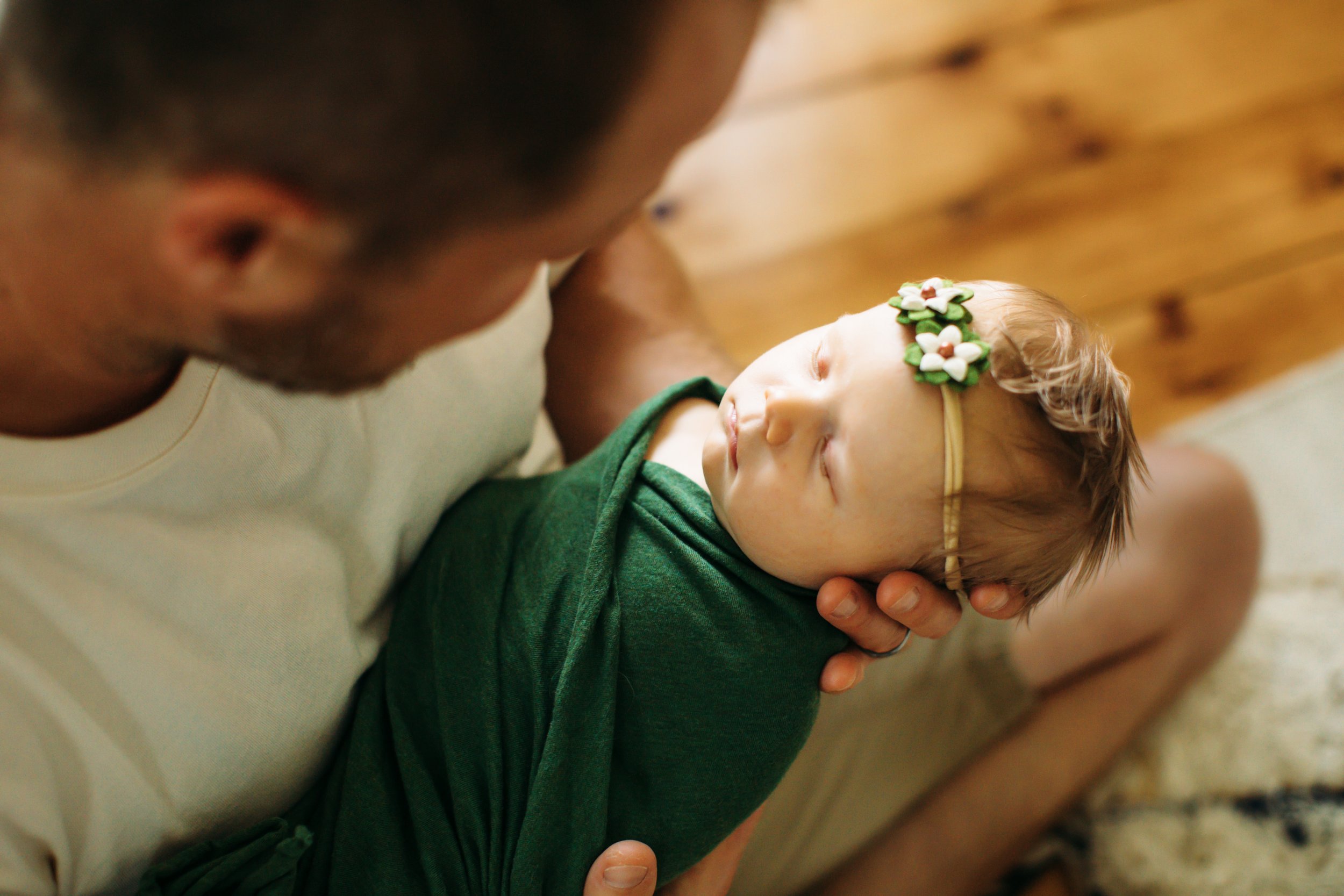  A newborn photo shoot at home will help keep your baby calm and happy. Teala Ward Photography image of a newborn baby girl being held by her dad while swaddled and wearing a flower headband #TealaWardPhotography #illinoisphotographer #princetonillin