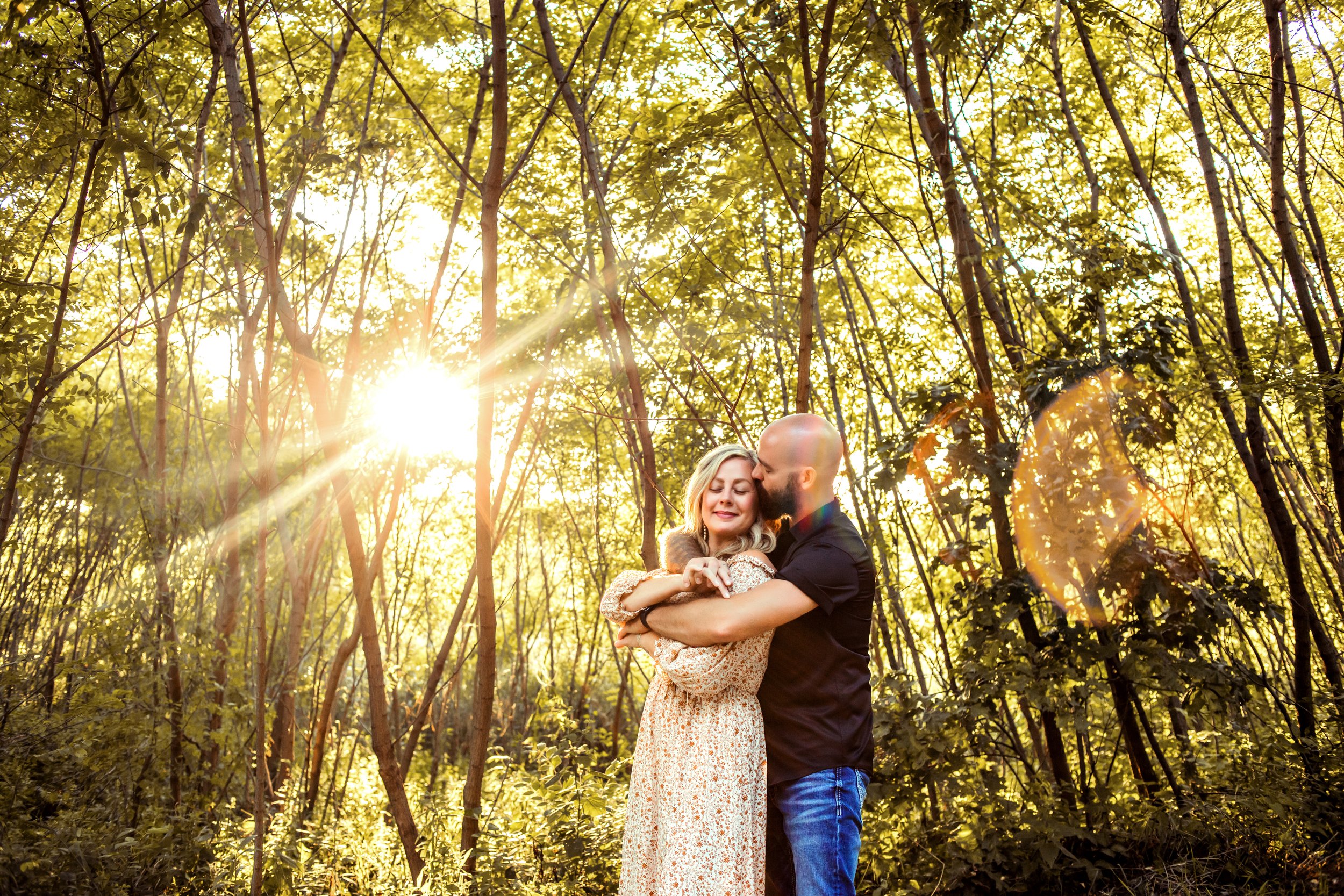  The sun shines through a forest illuminating a husband and wife hugging by Teala Ward Photography. Dynamic family pictures #TealaWardPhotography #IllinoisValleyPhotographer #midwestphotography #TealaWardFamilies #Illinoisfamilypictures 