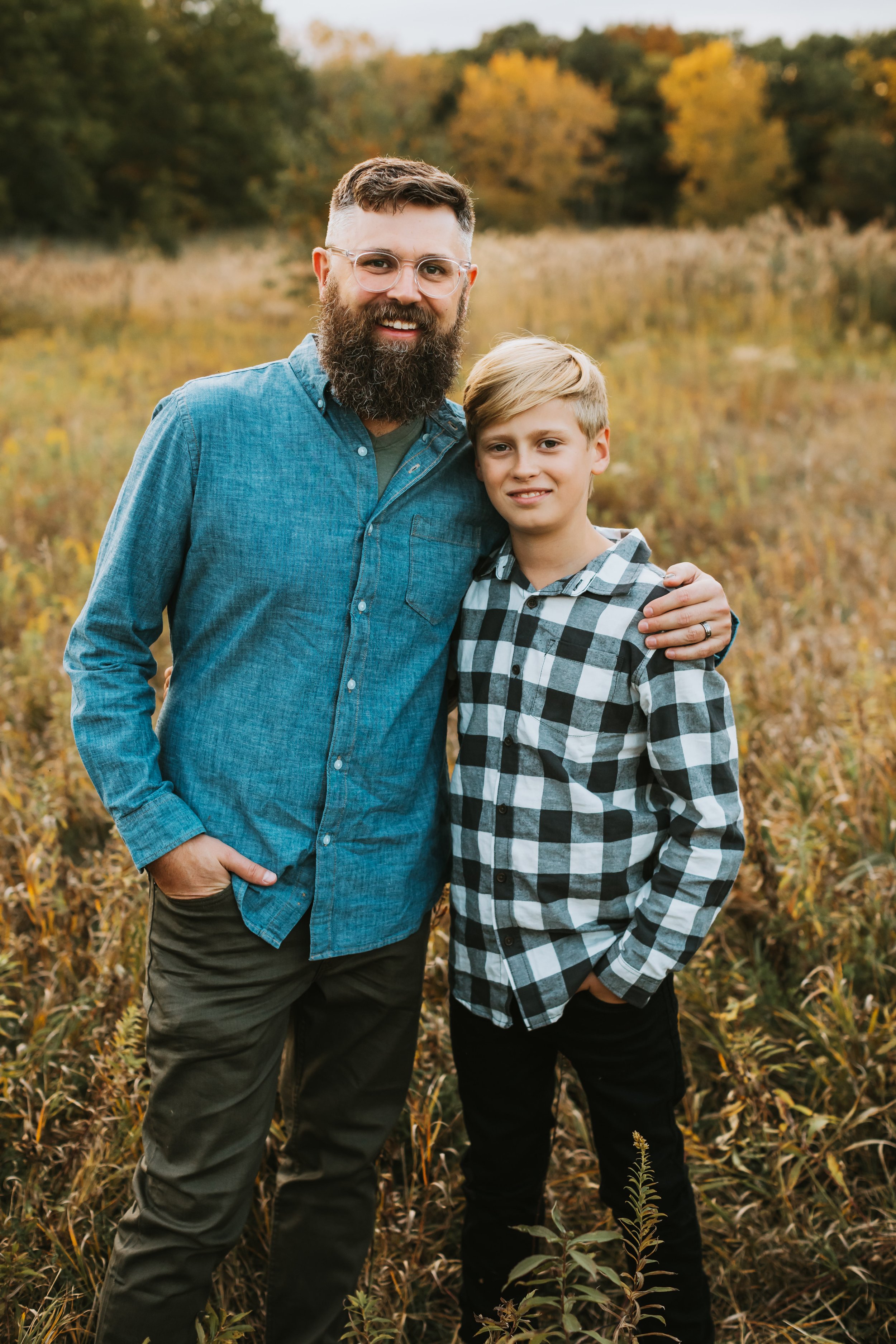  A hip father wearing a denim button-up and glasses smiles with his son in a plaid shirt by Teala Ward Photography. father son #TealaWardPhotography #TealaWardFamilies #IllinoisValleyfamilypics #fursibiling #Illinoisfamilyphotography #brother&amp;sis