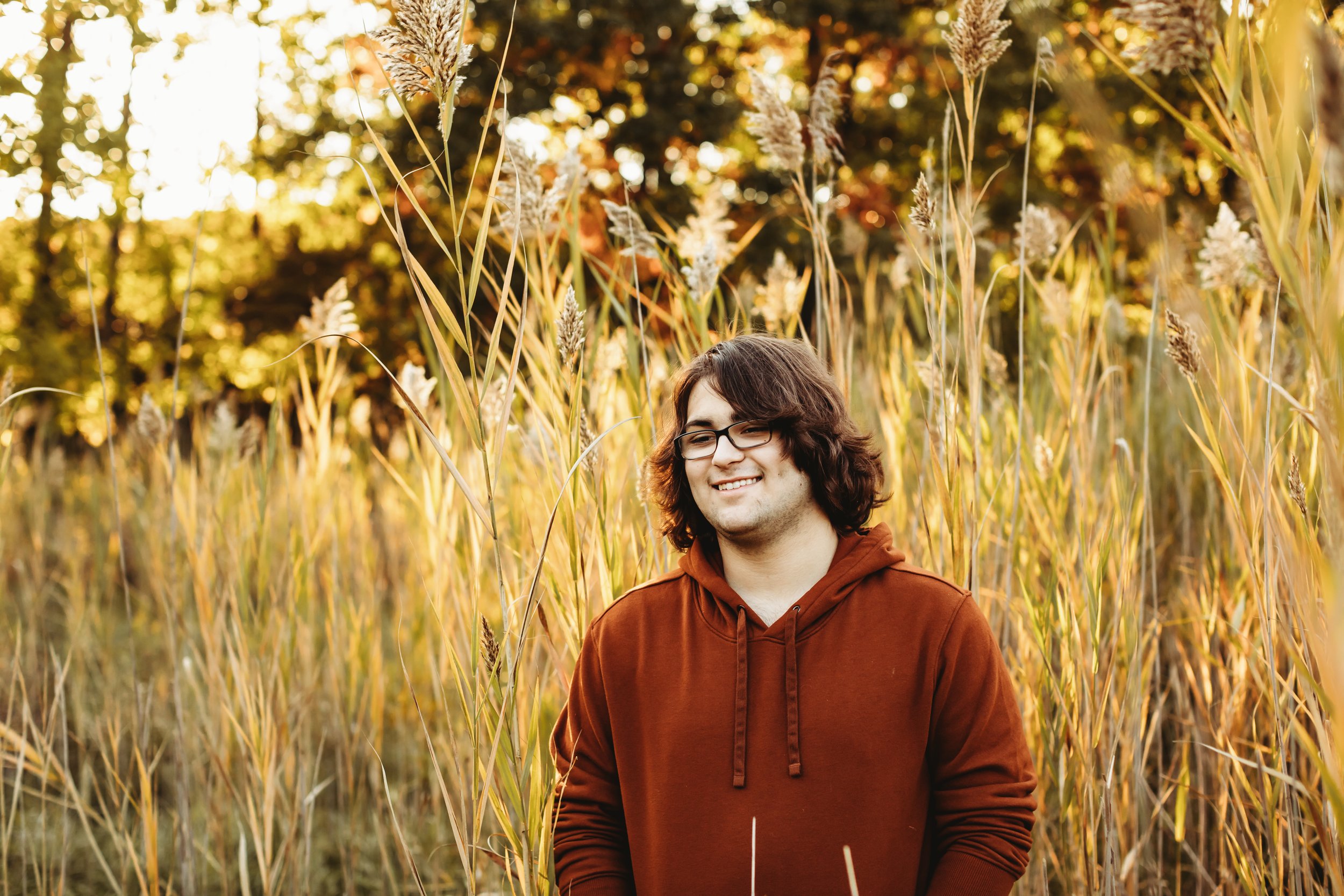  Portrait of teenage son smiling in yellow grass field wearing a rust-colored hoodie by Teala Ward Photography. rust colored outfit for teenage boy Illinois portraits #TealaWardPhotography #TealaWardFamilies #Outdoorfamilyportraits #IllinoisPhotograp