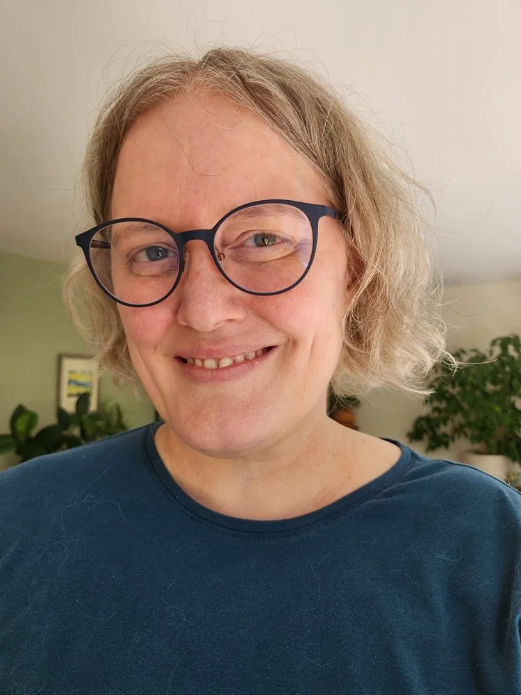 A middle-aged white female with chin length wavy light-colored hair and dark blue glasses smiling at the camera.