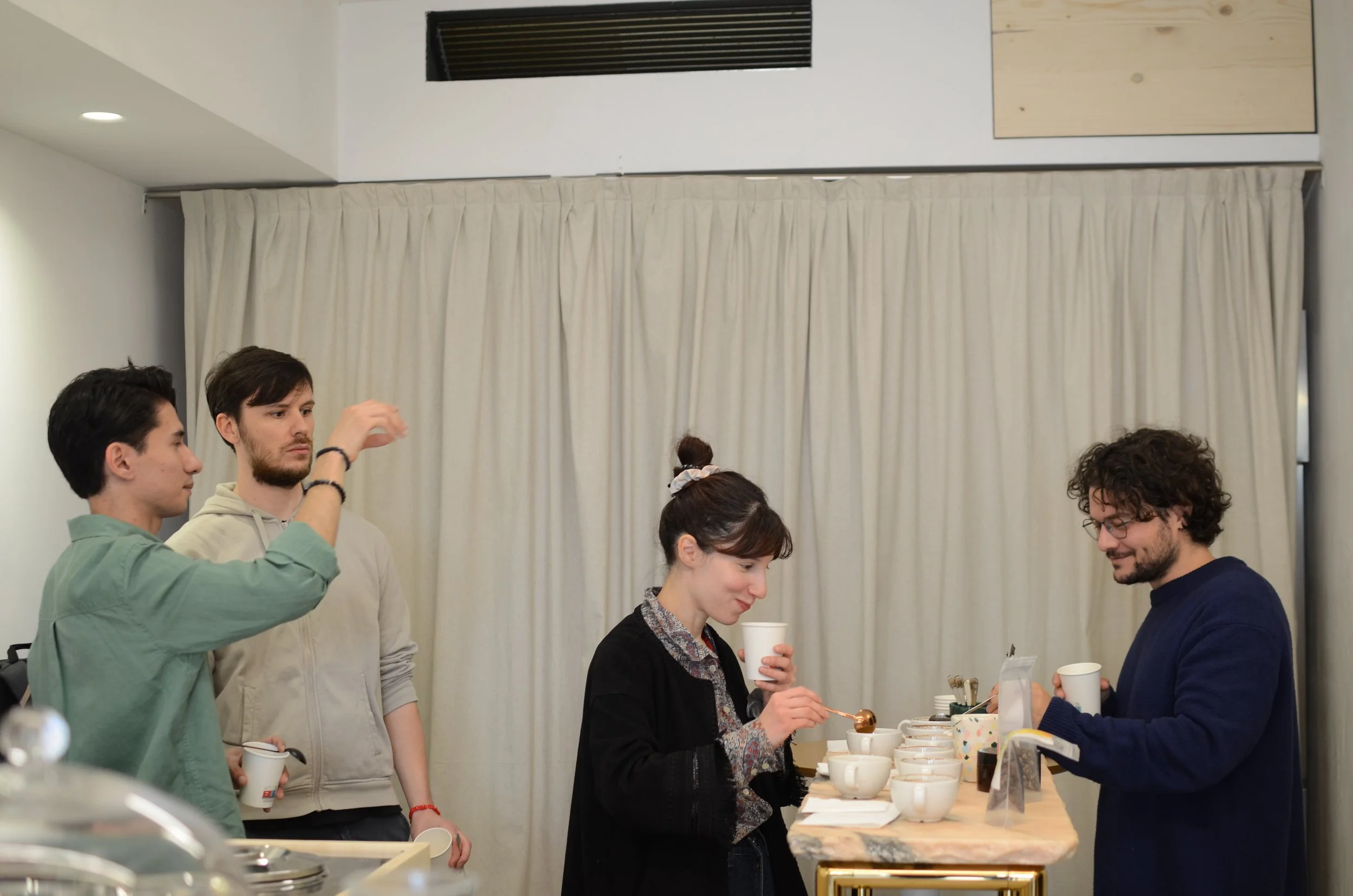 Four young adults stand at a table with cups and bowls, engaging in a conversation in a room with white curtains.