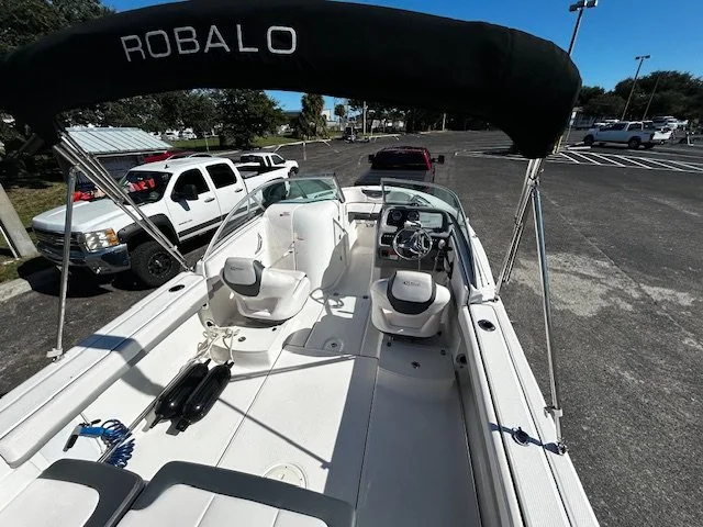 View of a white boat with a black Bimini top labeled Robalo, parked in an outdoor lot with other vehicles and parking spaces visible in the background.