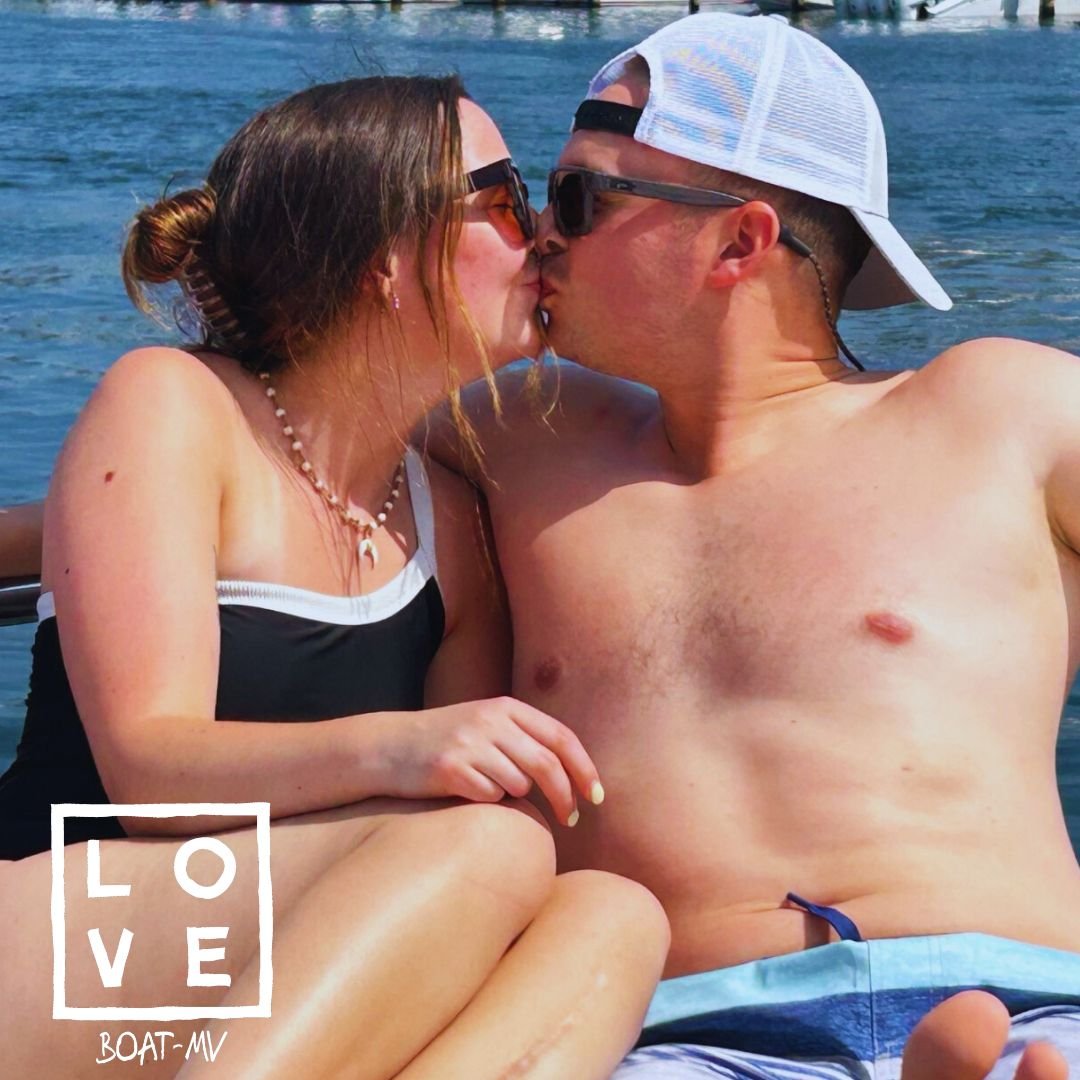 A couple kissing on a boat, with water in the background, both wearing sunglasses, and the man wearing a white cap backwards.