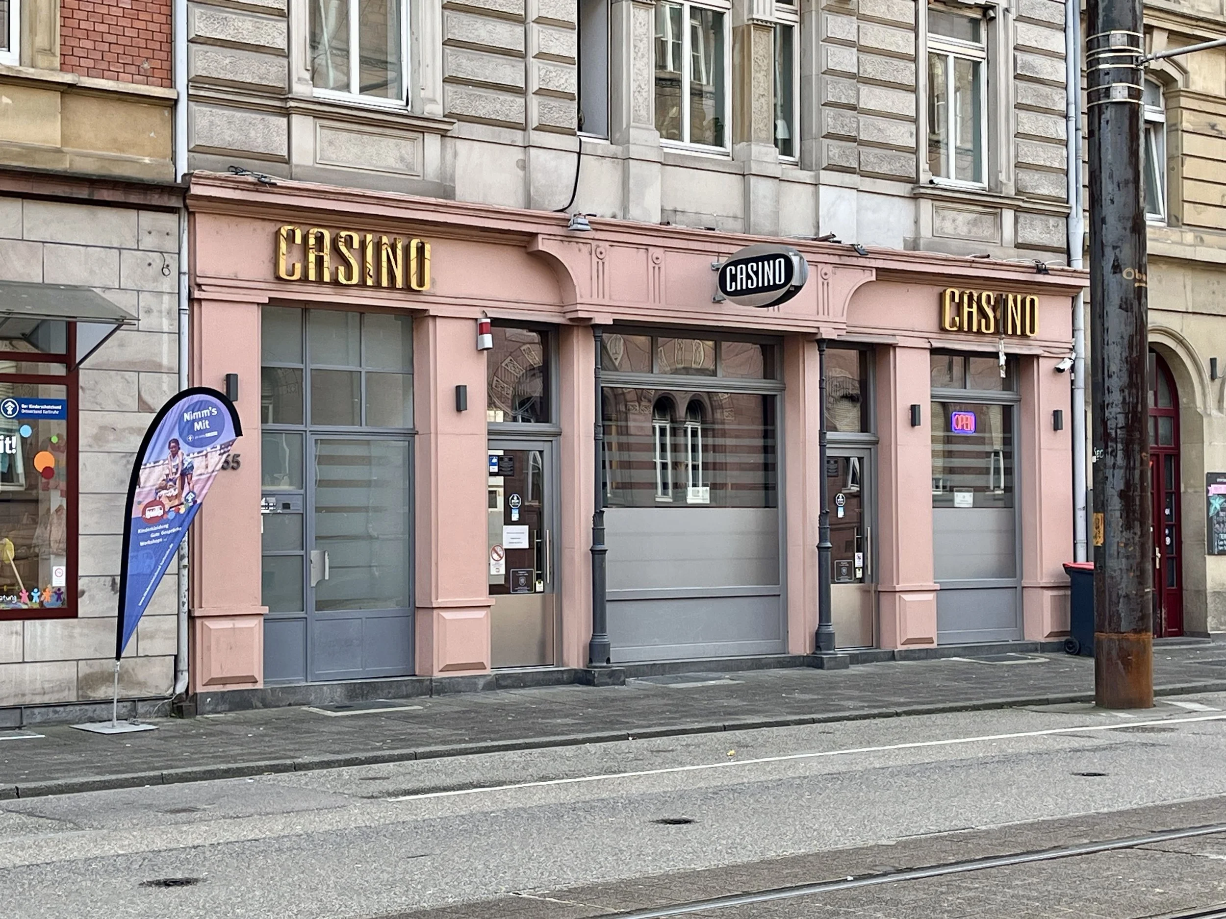 Ein historisches Gebäude mit rotem Sandstein in einer städtischen Umgebung, umgeben von modernen Gebäuden, mit Autos und Straßenbahnschienen im Vordergrund. Nähe KIT