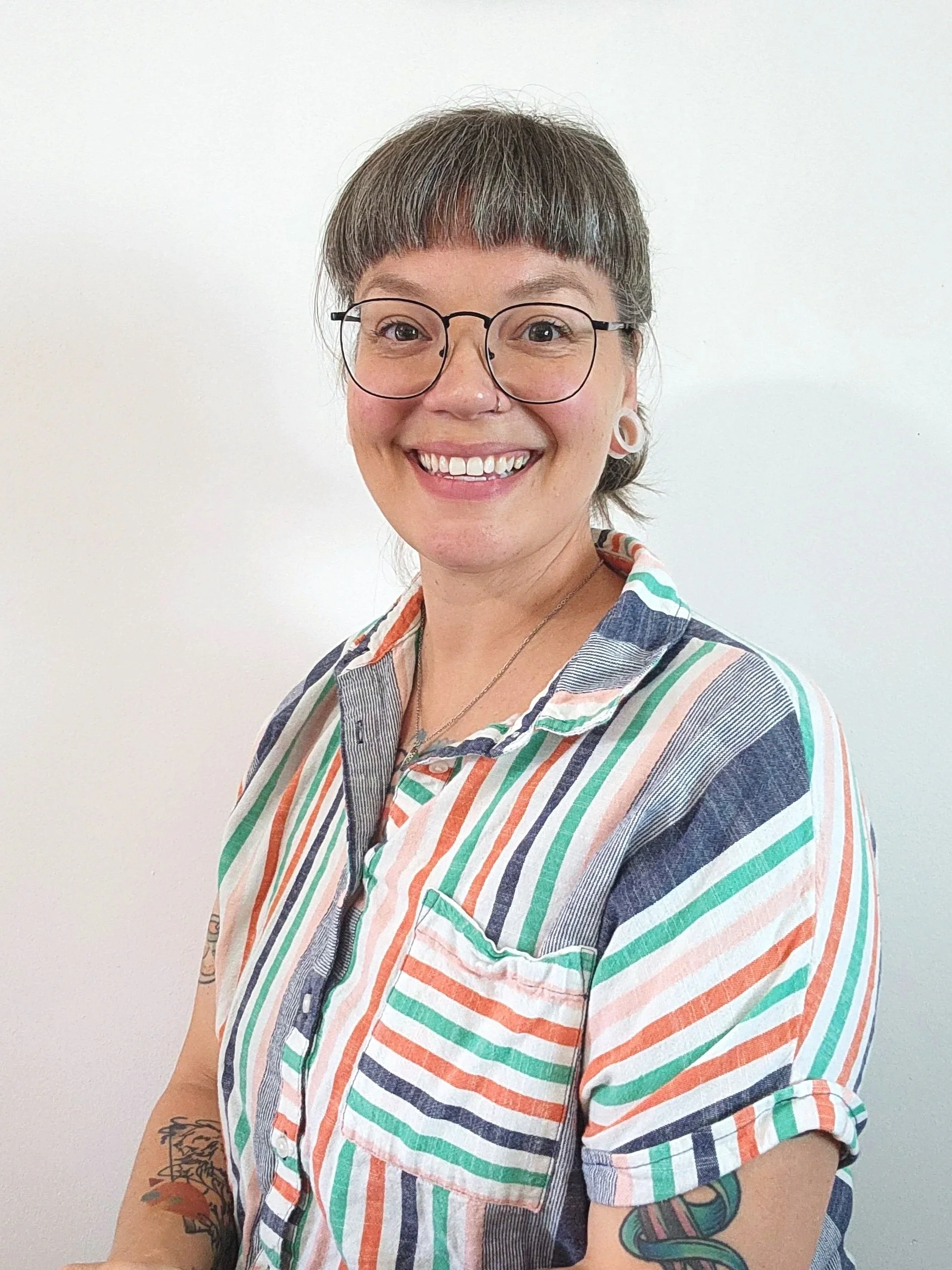A person with short, styled hair, wearing large black glasses, earrings, and a black top, smiling with tattoos visible on their chest.