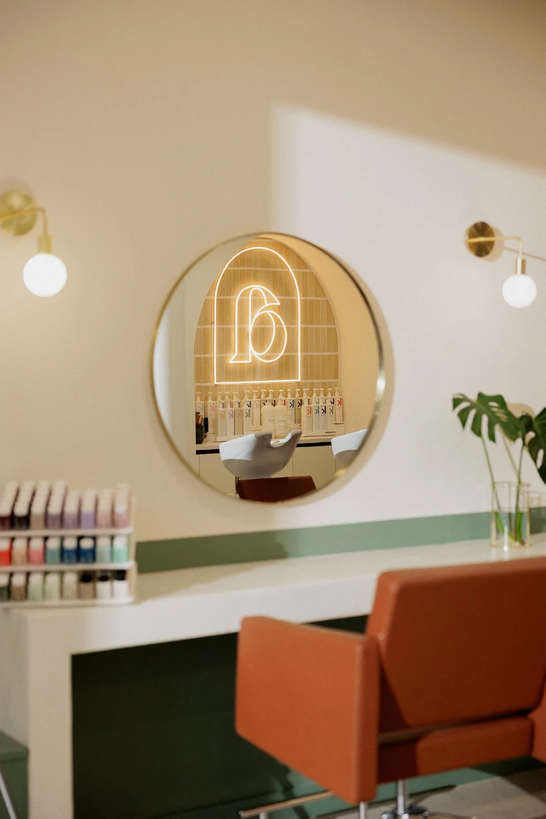 Chaise de salon de coiffure élégante avec miroir rond, étagère avec shampoings miniatures, plante verte dans un vase, et deux lampes murales dans une pièce lumineuse.