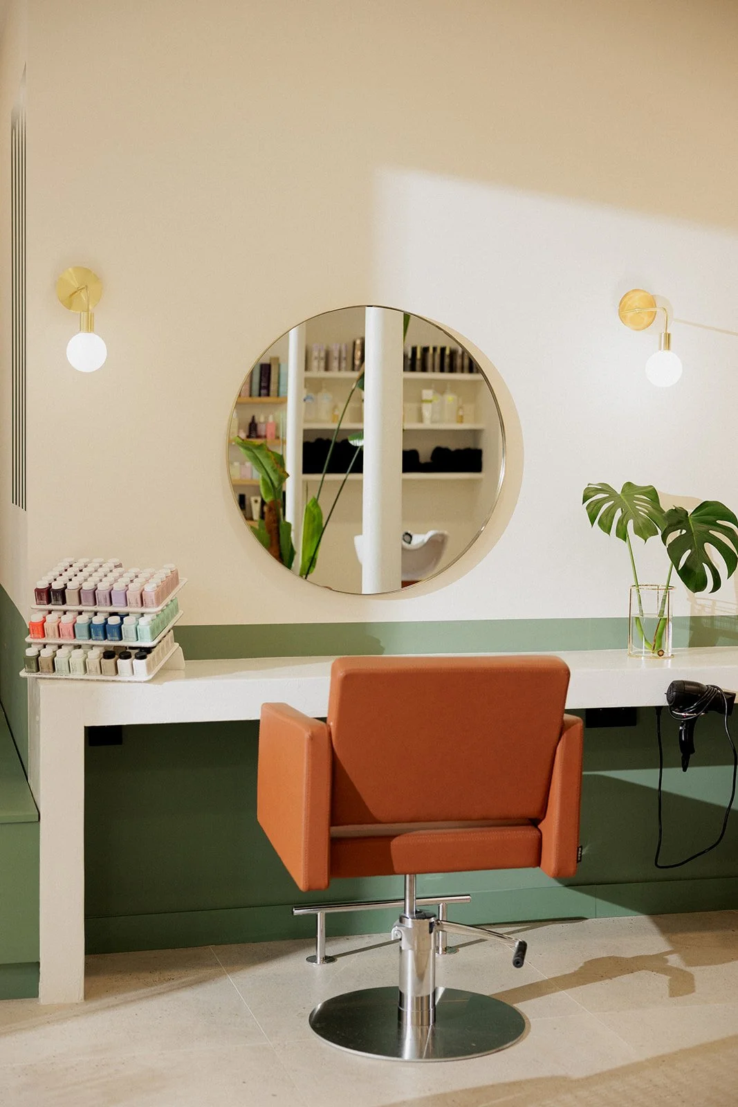 Une chaise de coiffure en cuir marron devant un miroir rond, une plante verte, une étagère avec des produits, deux luminaires