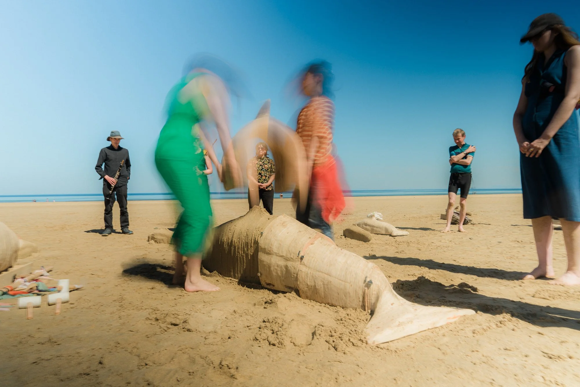 Artist Sam Gare and choreographer Aya Kobayashi lift a section of the whale mould of the sand whale created by the community, while musicians look on and play
