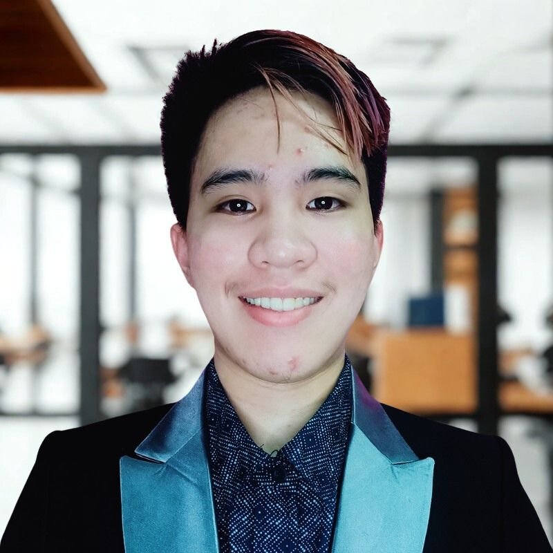 Kirby, Administrative Assistant at Touch by the Angel, smiling with short partially dyed hair in a black blazer and a blurred office background.