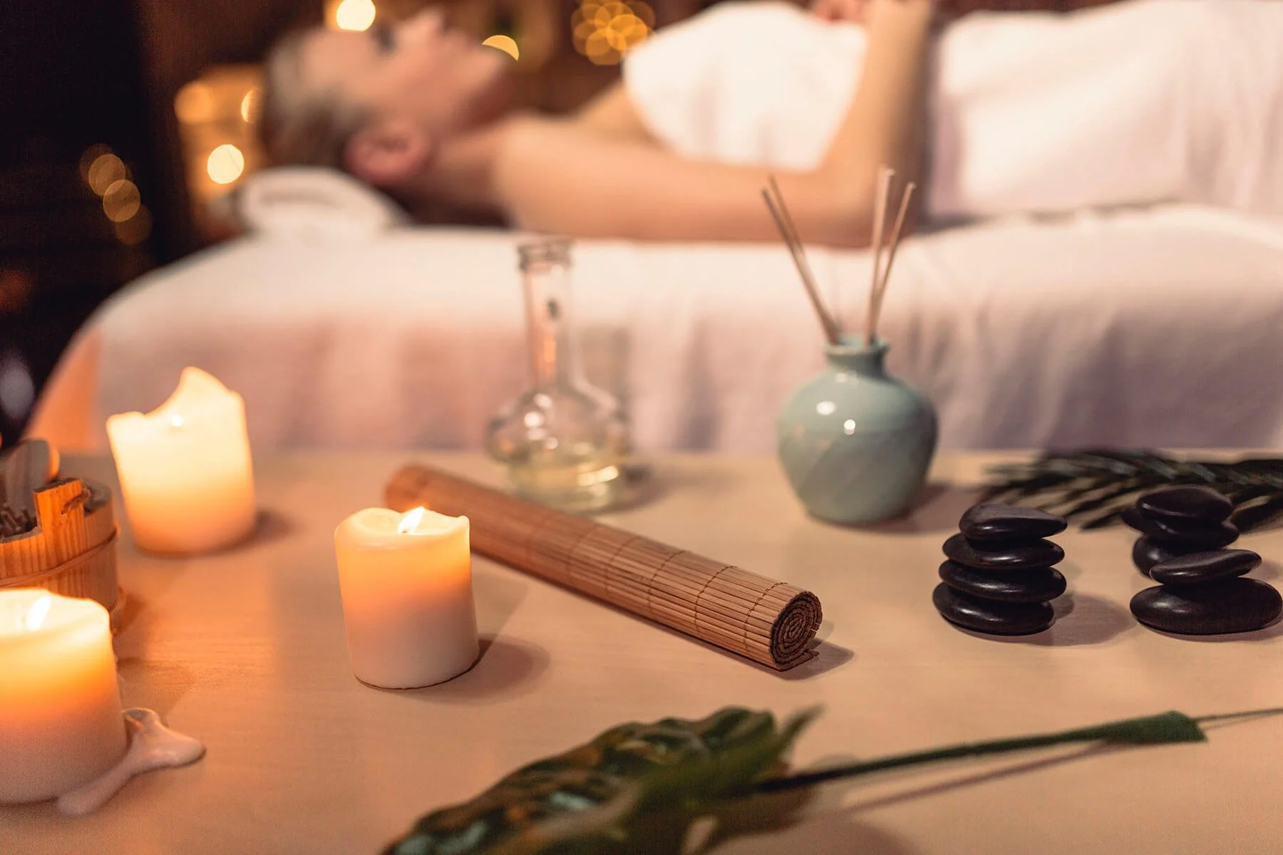 A person relaxing on a bed in the background with spa and massage items on a table in the foreground, including candles, a rolled towel, stones, bamboo sticks, and incense.
