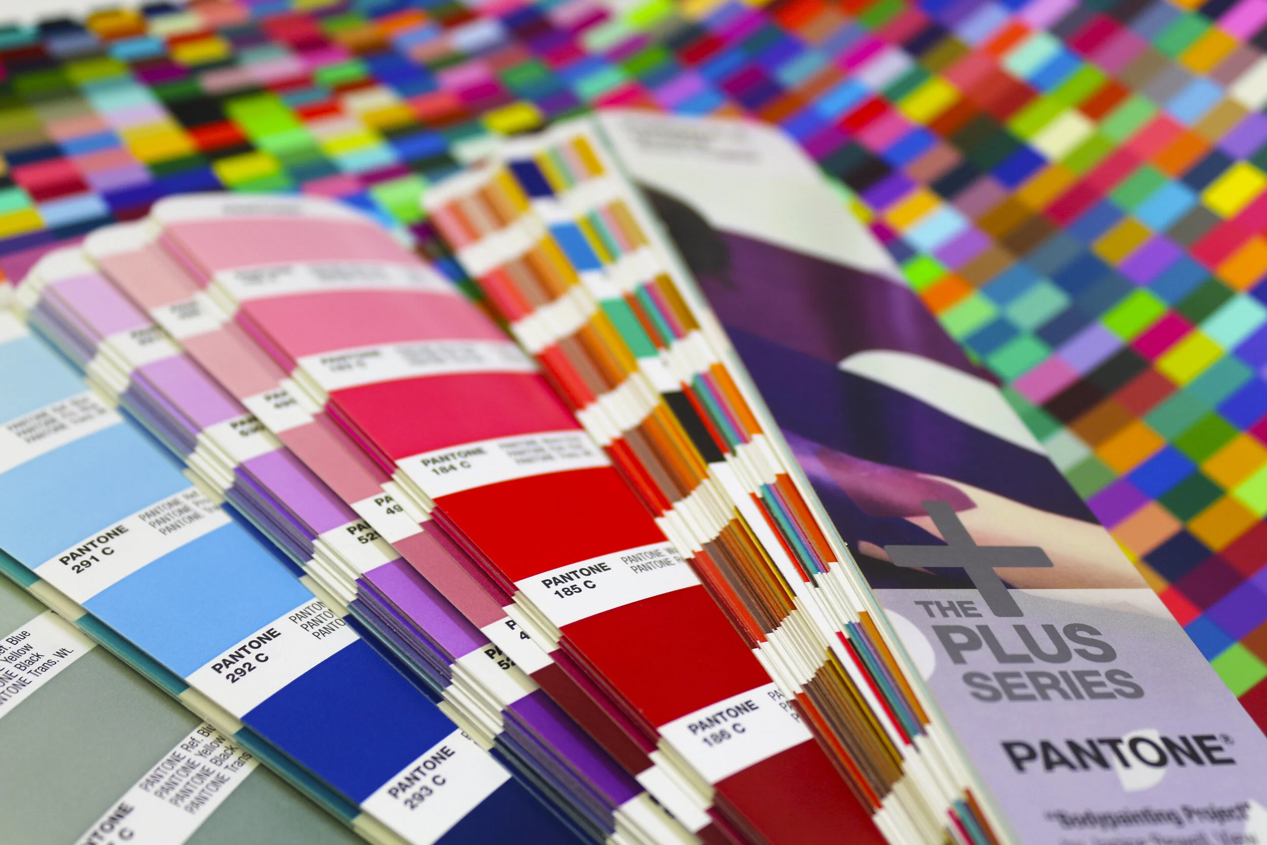 Color swatch fan decks with Pantone color samples and a promotional banner for the Pantone Plus Series on a multicolored mosaic background.