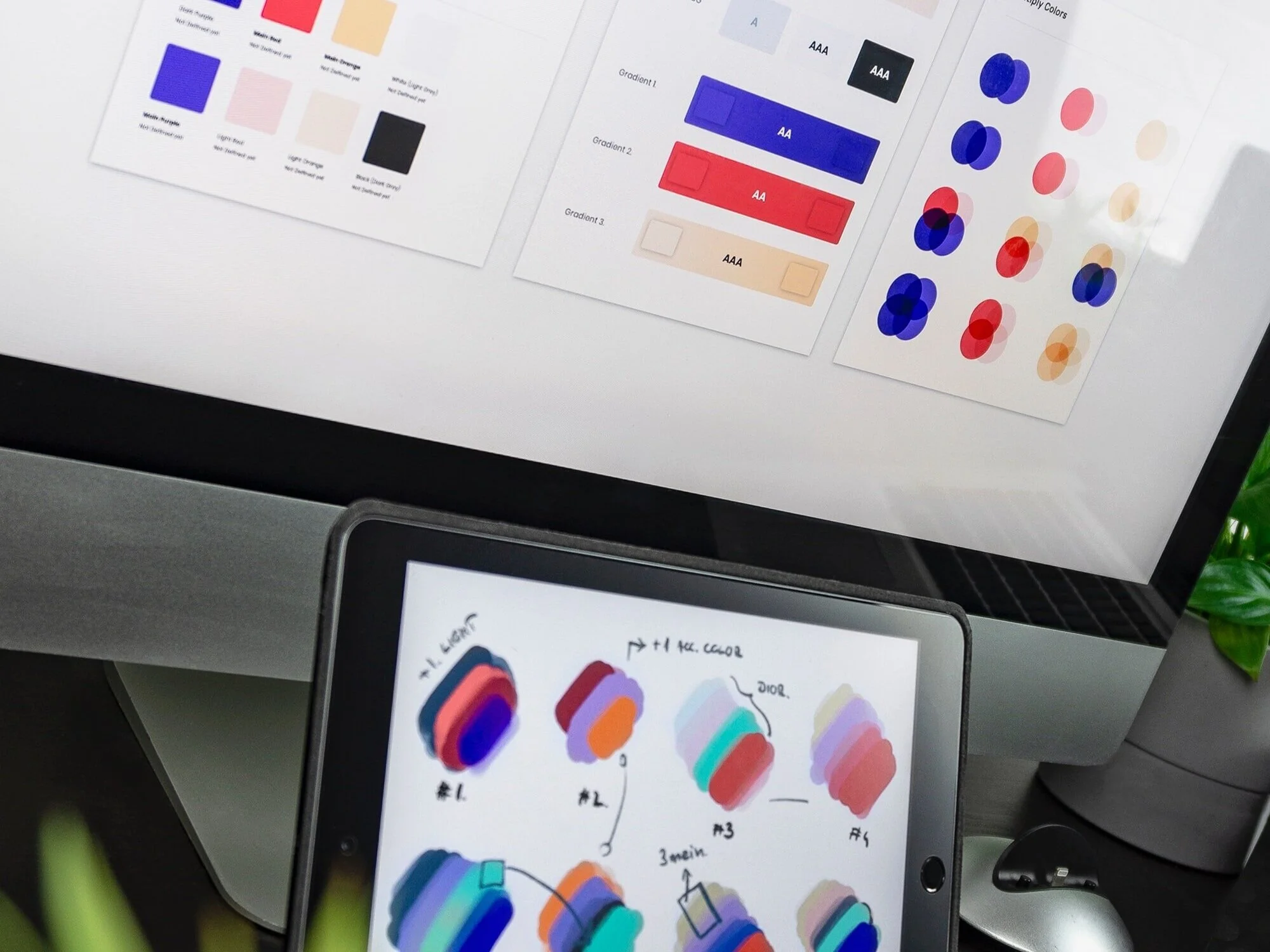 Color palettes and design documents on a computer monitor and tablet, with a potted plant partially visible on the right to demonstrate the importance of color management.