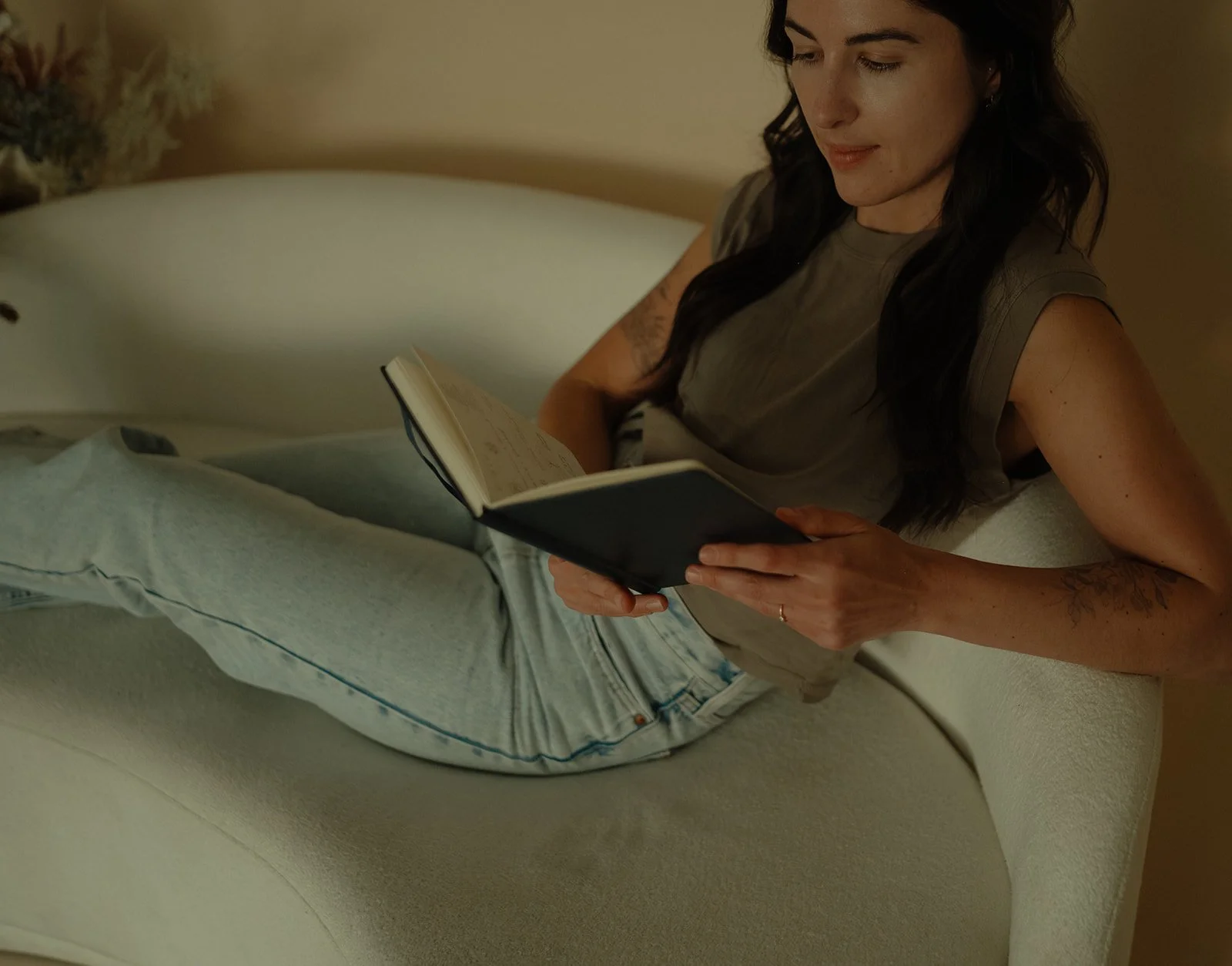 A woman with dark hair reading a book while lying on a cream-colored sofa in her studio in Central Oregon.