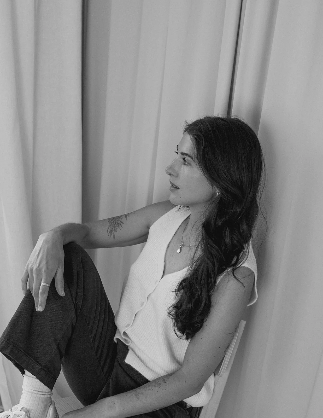 A black and white branding photo of a woman with long, wavy hair sitting on the floor against a wall. She is wearing a sleeveless shoes, with a tattoo on her upper left arm and a necklace.